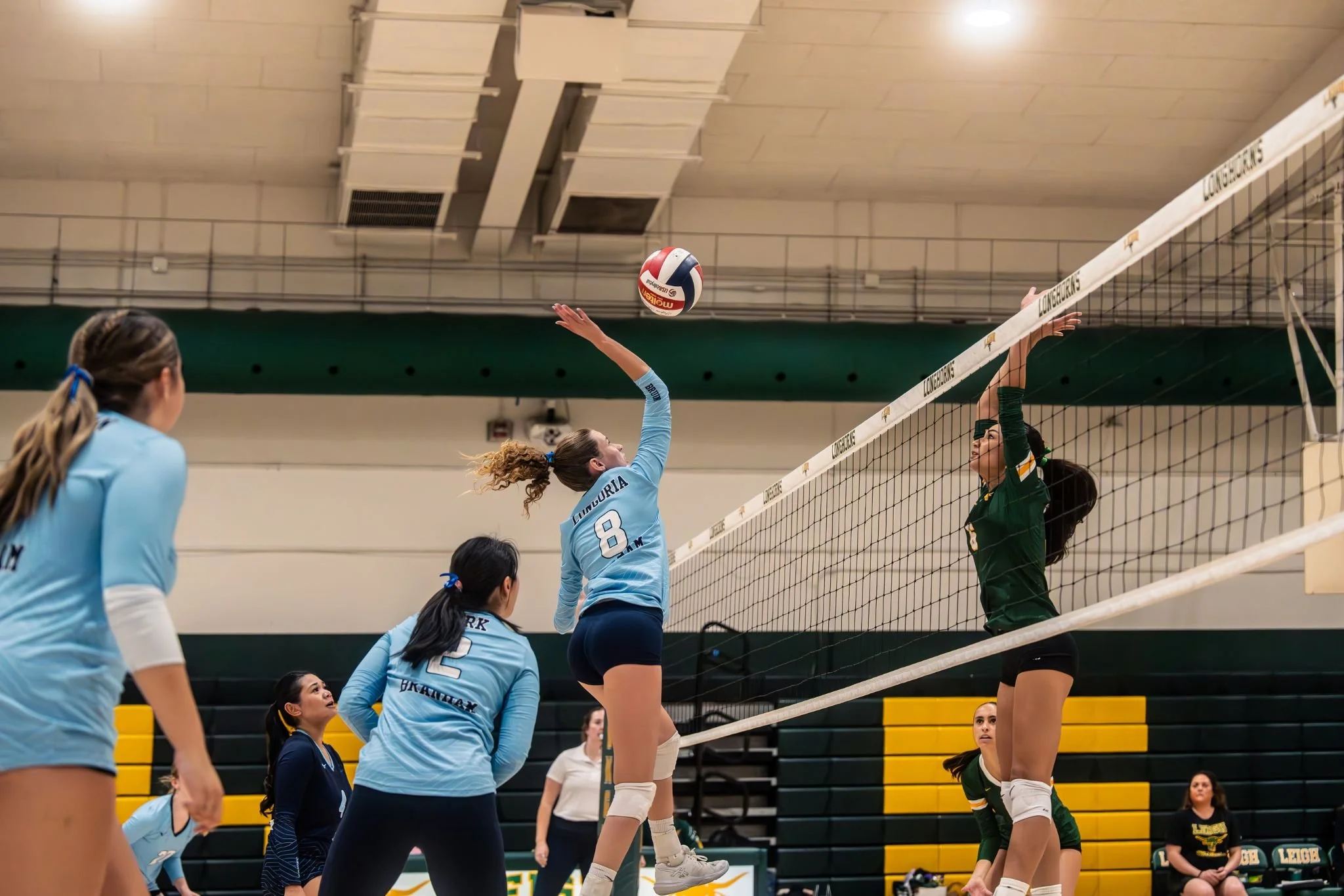 Female volleyball players on an indoor court during a game, with some attempting to block a spike at the net.