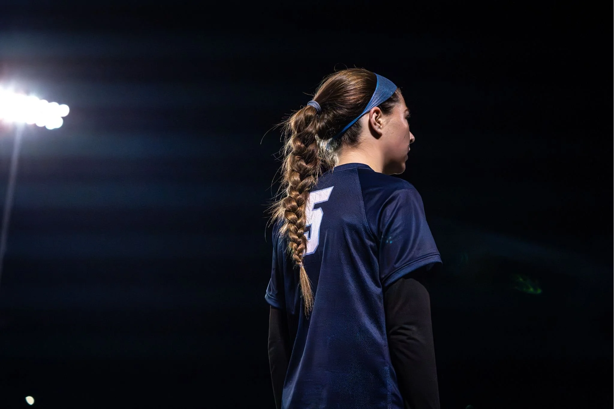 Profile view of a female athlete with braided hair, wearing a blue sports headband and jersey, standing under stadium lights at night.