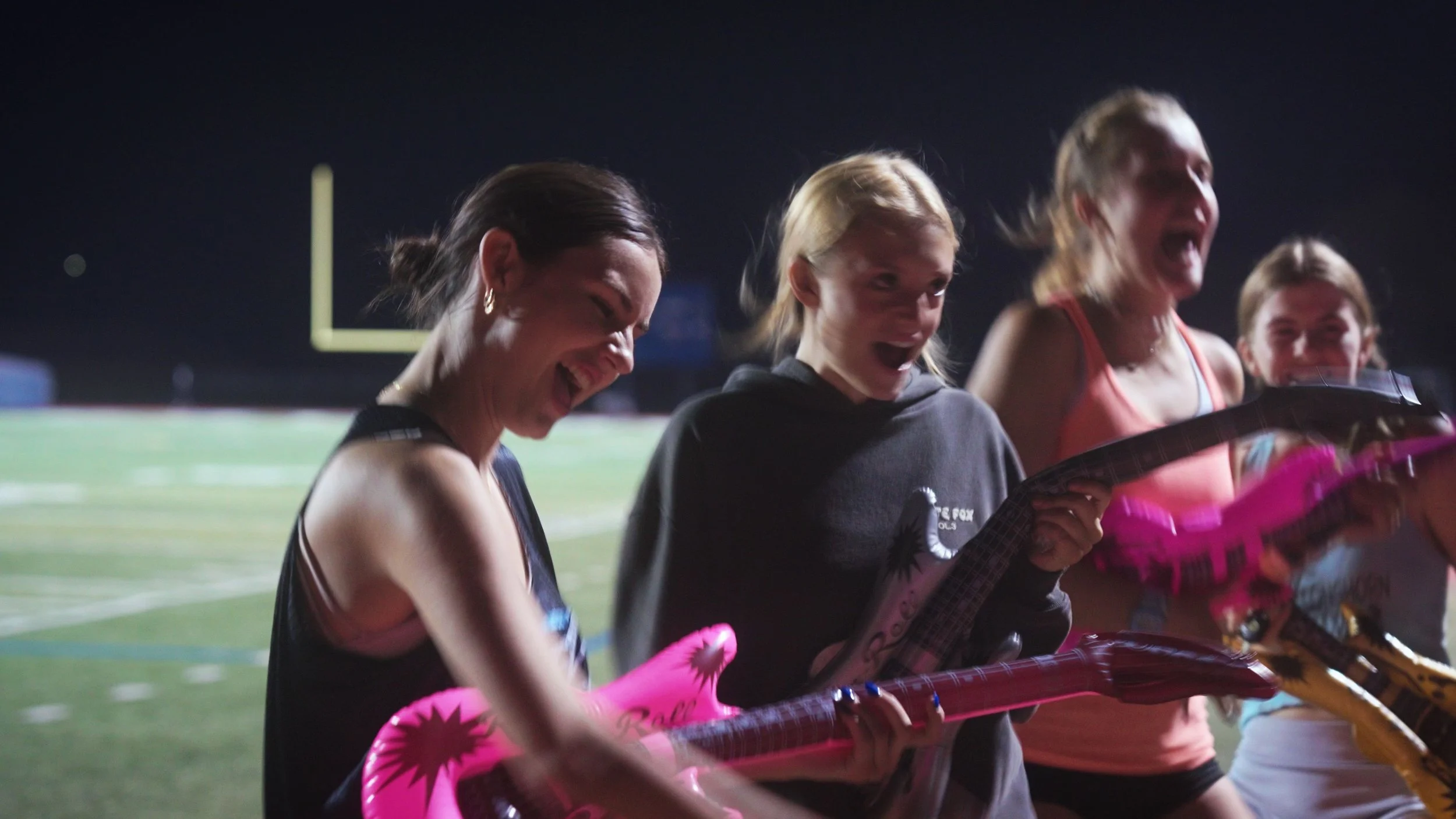 Four women standing on a football field at night, smiling and holding pink and purple foam instruments, celebrating.