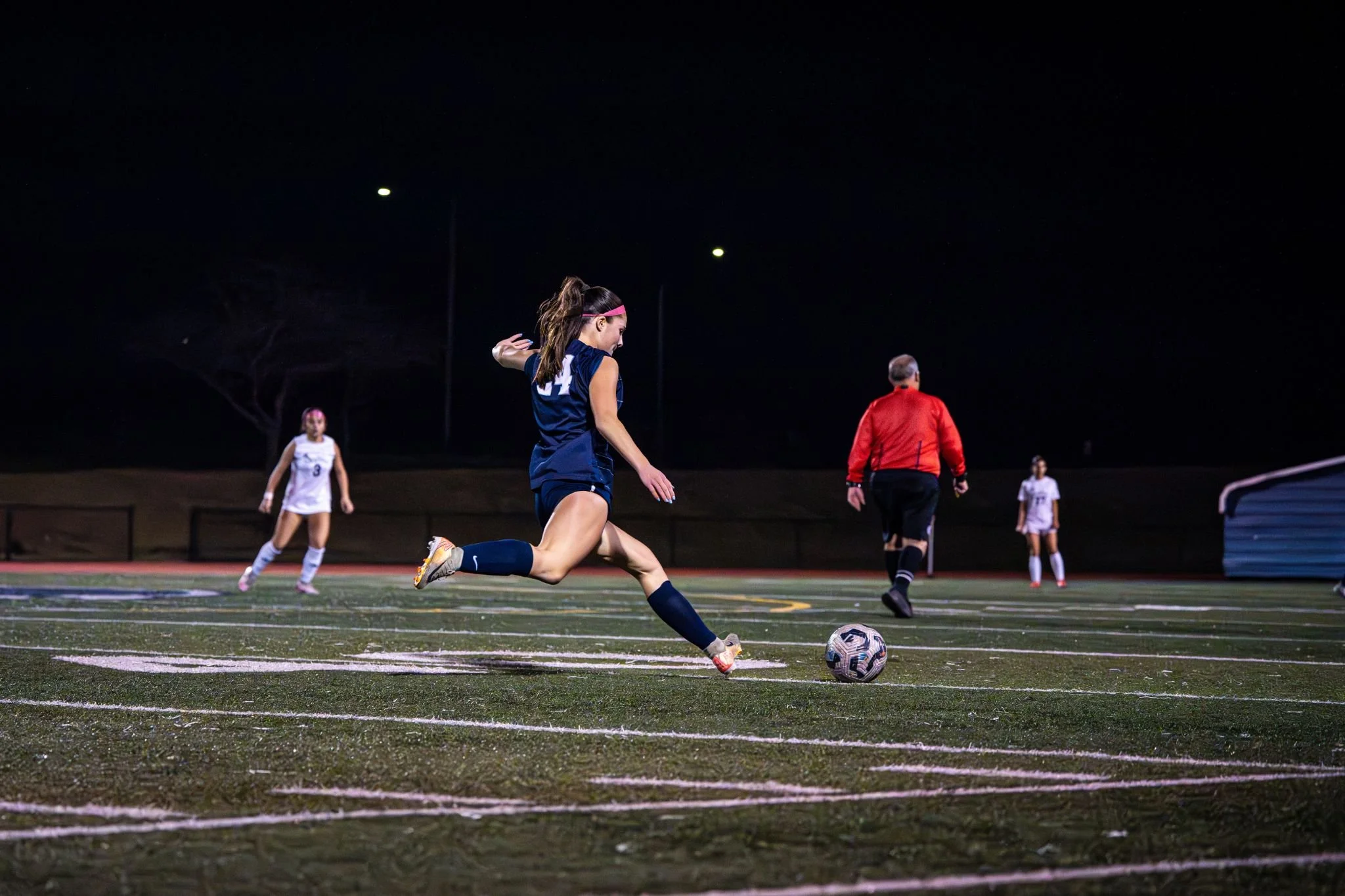 Nighttime soccer game with a female player in a blue uniform kicking the ball, surrounded by other players and a referee on a well-lit field.
