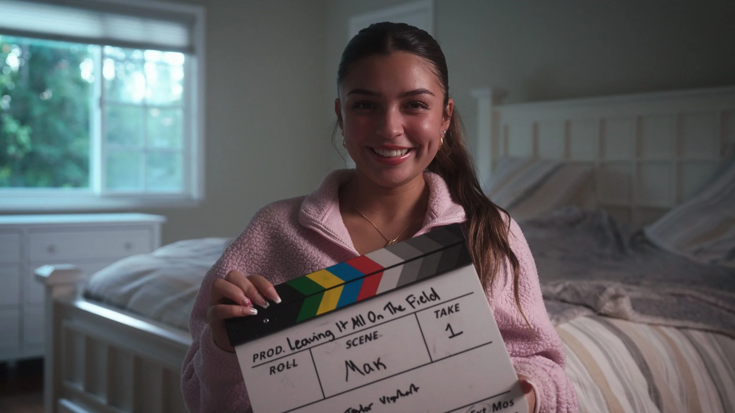 A smiling young woman with brown hair tied back, wearing a pink fleece jacket, holding a film slate in a bedroom with a bed and a window with greenery outside.