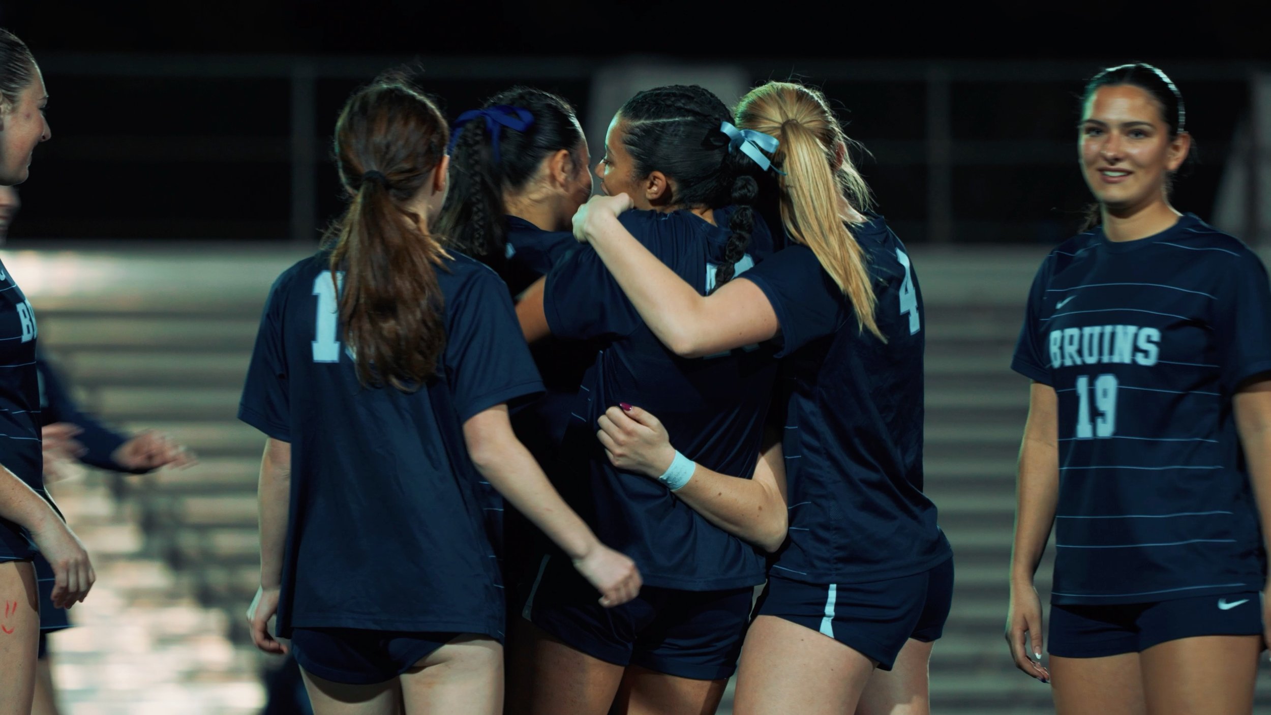Group of young women in navy blue sports jerseys celebrating on a sports field at night.