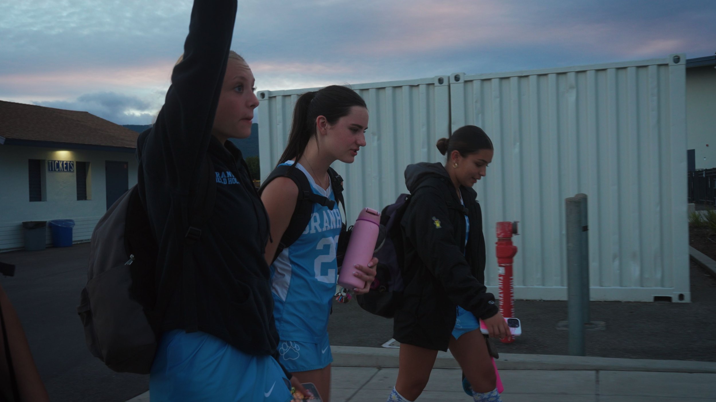 Three young women in athletic clothing walking outdoors during dusk, each carrying a backpack and water bottle.