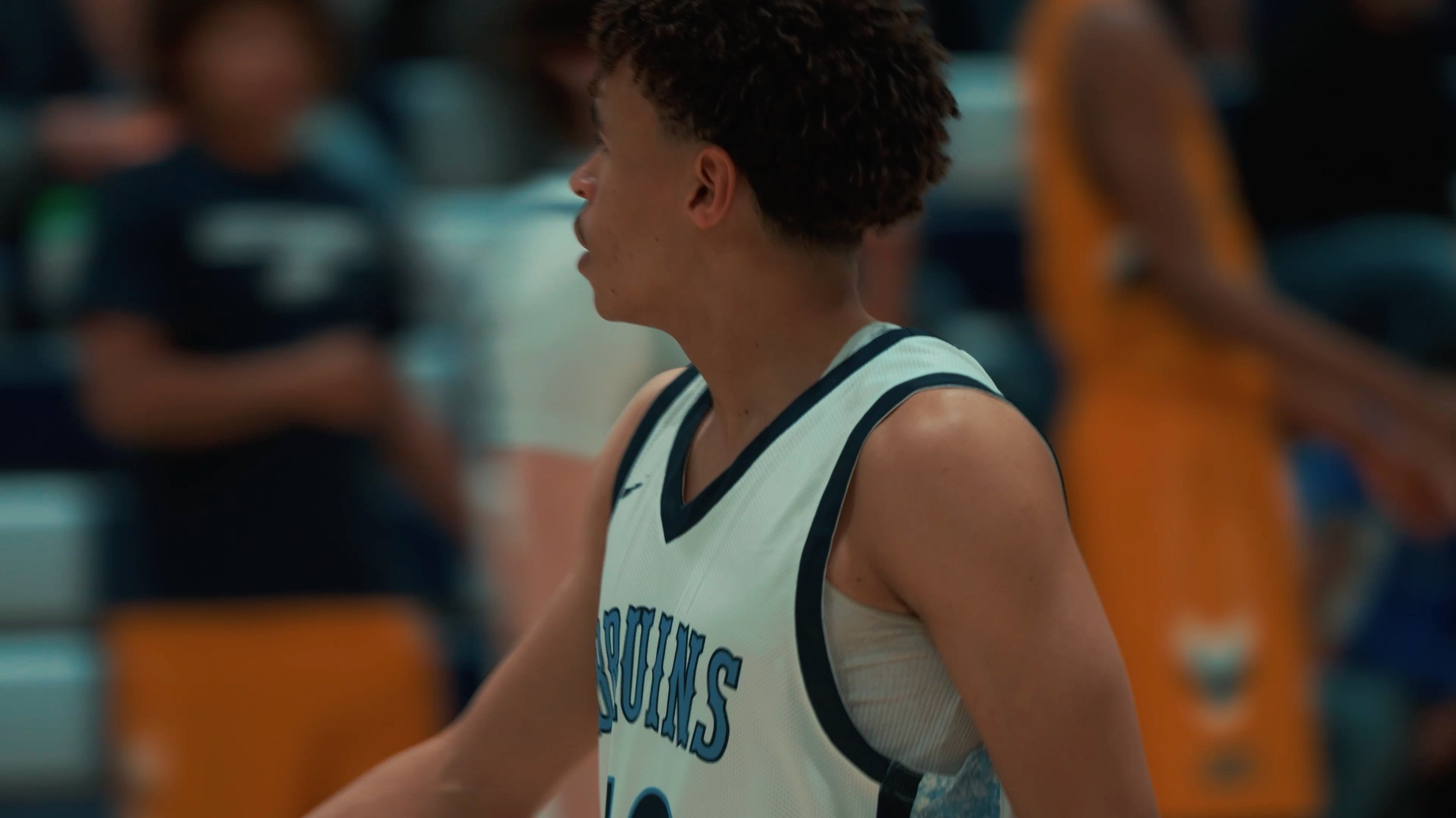 A young male basketball player with curly hair wearing a white and blue basketball jersey, standing on a basketball court with blurred players in the background.