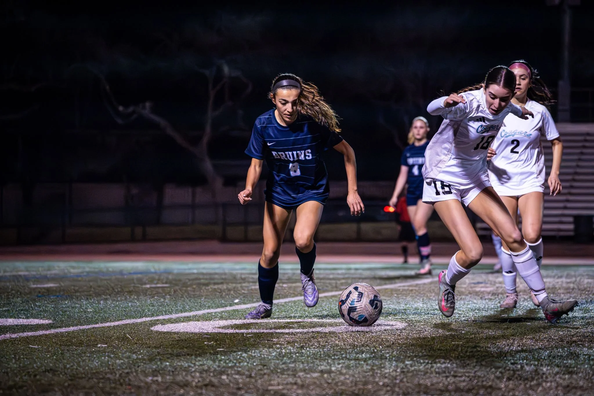 Female soccer players competing for the ball during a game at night.