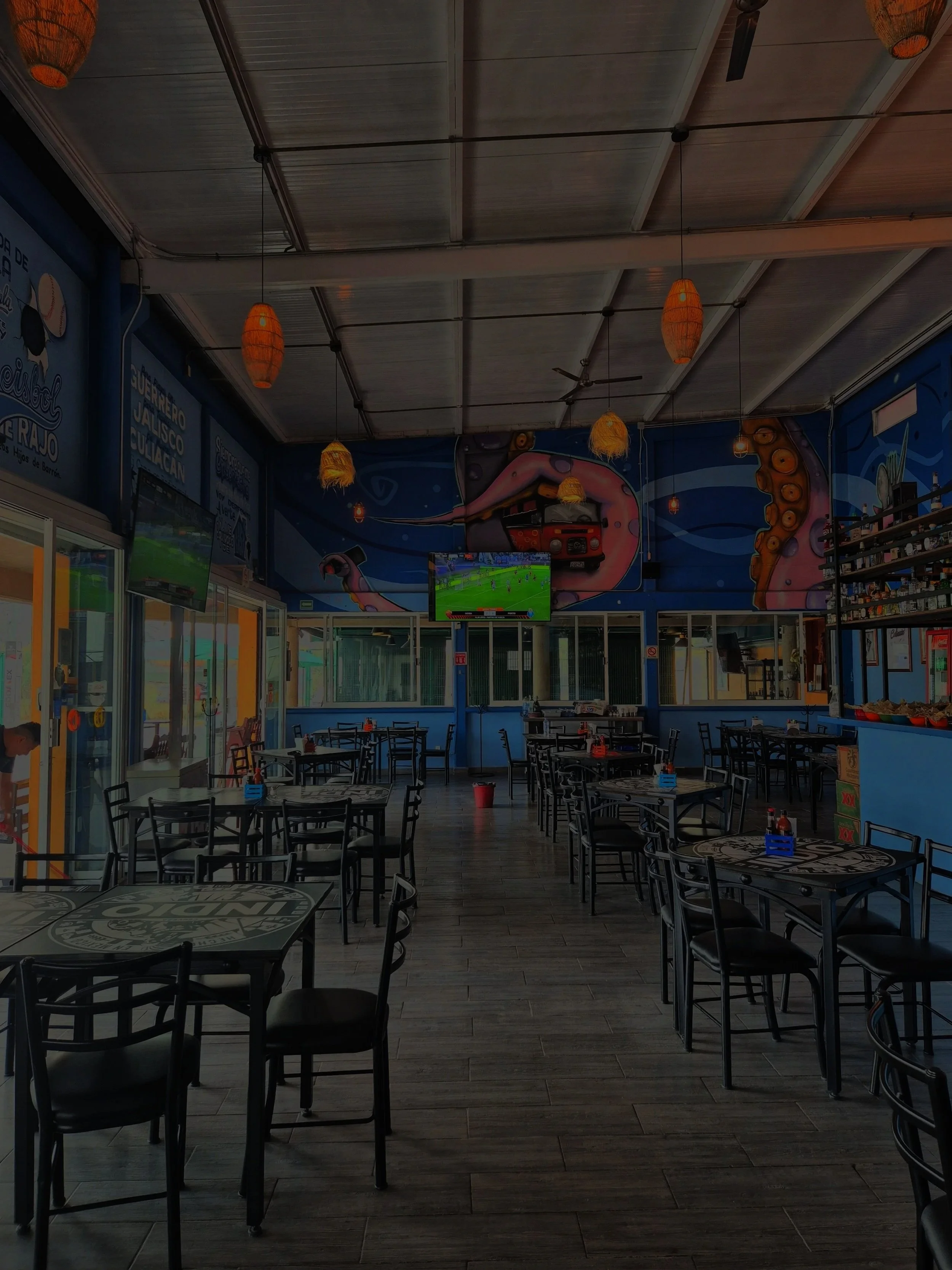 Interior de un restaurante con mesas y sillas, decorado con pintura de un pulpo y un camión en la pared, pantallas de televisión y lámparas de mimbre colgantes.