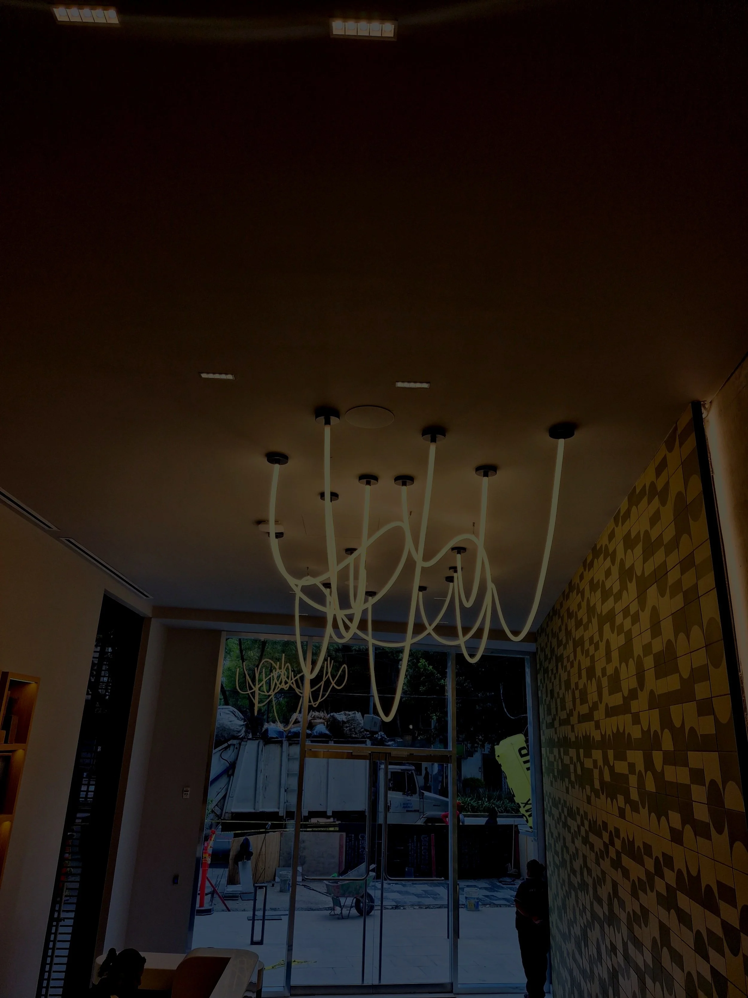 Interior de un establecimiento en construcción, con techo, lámparas, muro decorativo y ventanas grandes, vista a la calle con árboles y vehículos.
