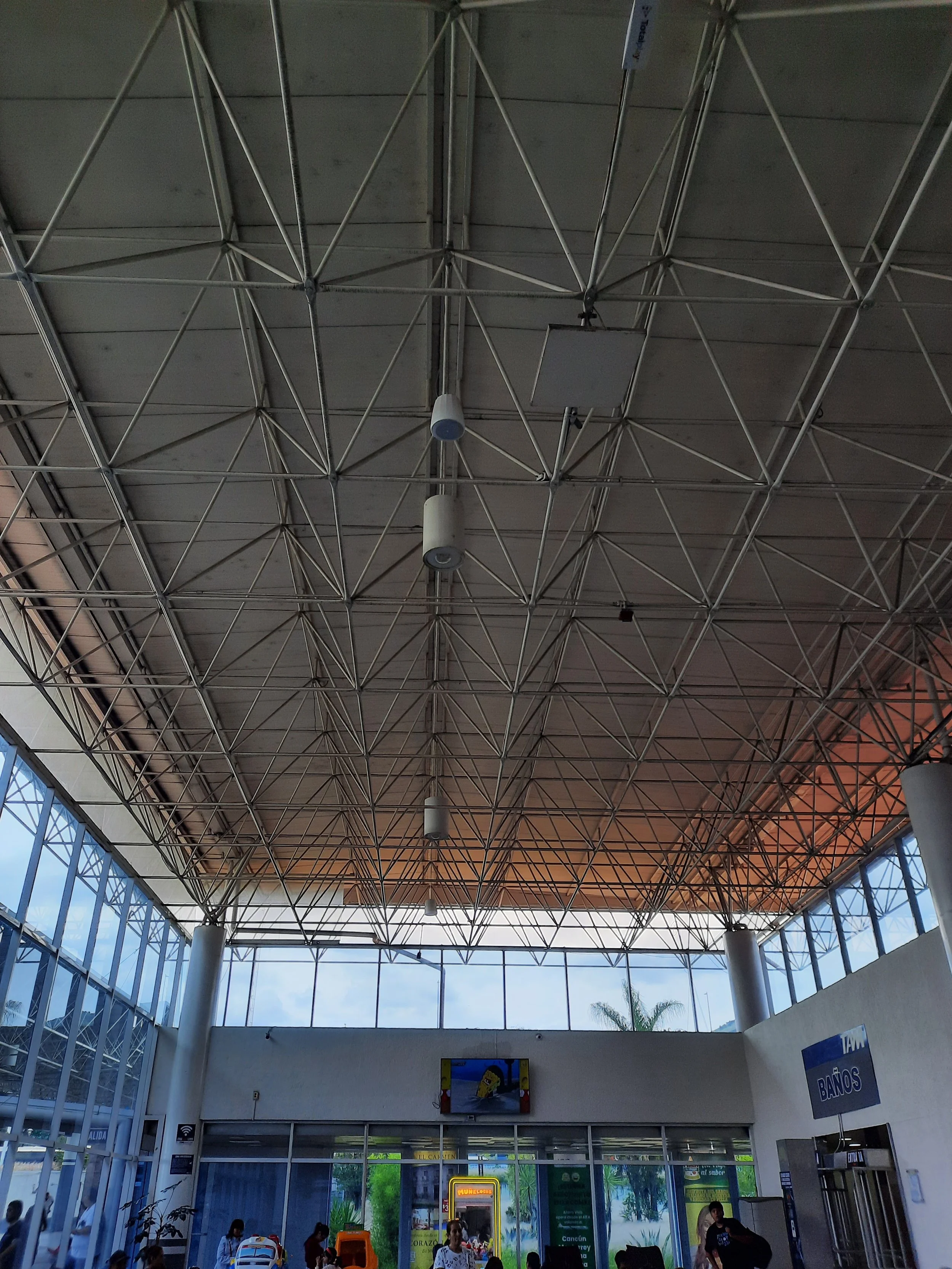 Interior de un aeropuerto con estructura de techos metálicos y ventanas grandes. Hay personas esperando, algunos con maletas, y un cartel de baños visible a la derecha.