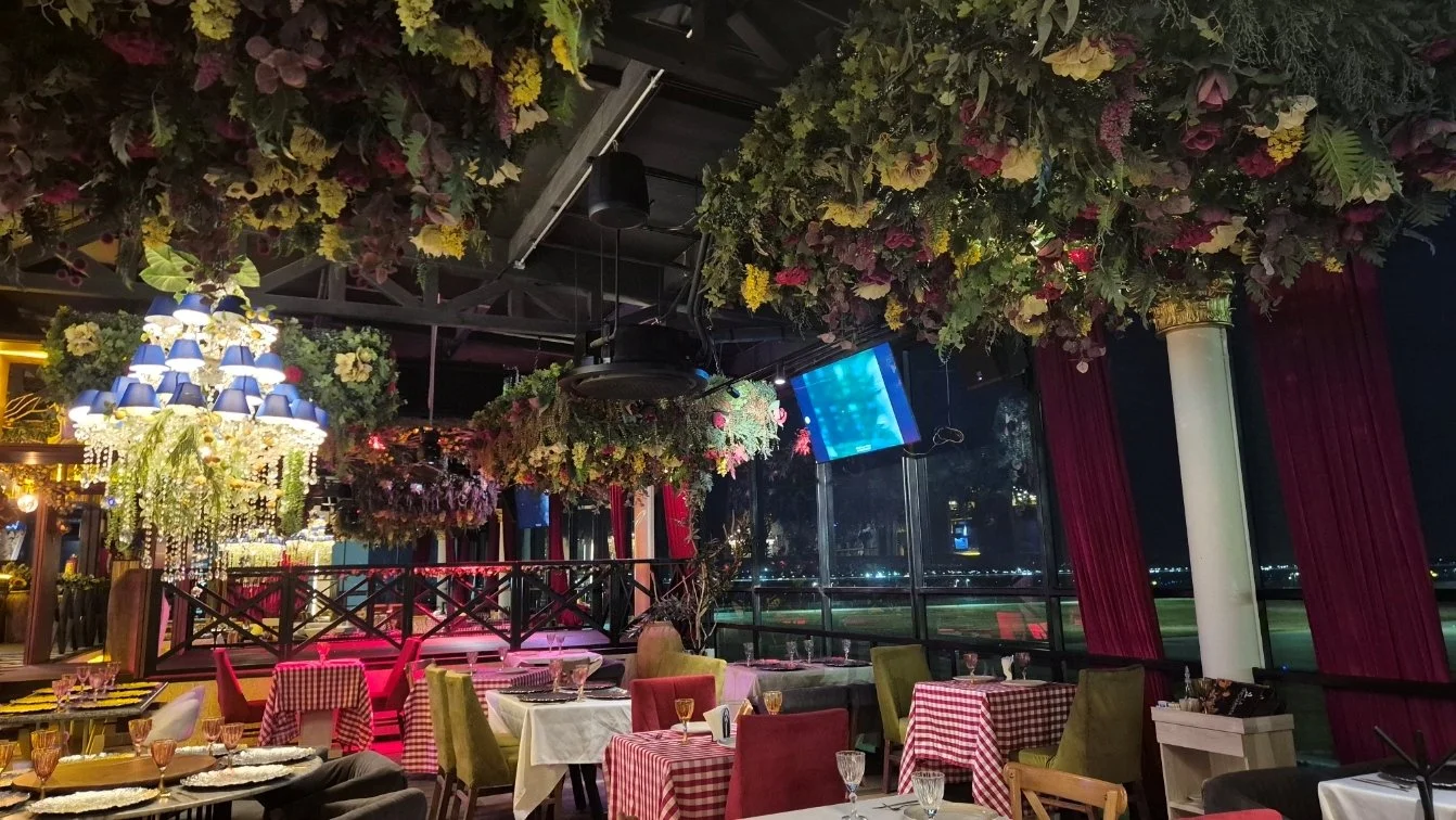 Interior de un restaurante elegante con mesas cubiertas con manteles de diferentes colores y cristalería, decoraciones florales en el techo y alrededor, y una vista nocturna a través de ventanas grandes.