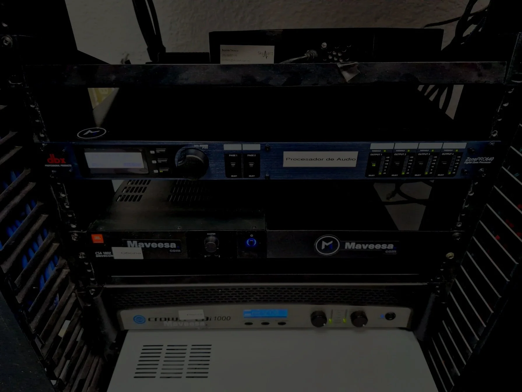 Rack de equipos de sonido, incluyendo un procesador de audio y otros dispositivos electrónicos, en un entorno de oficina o estudio.