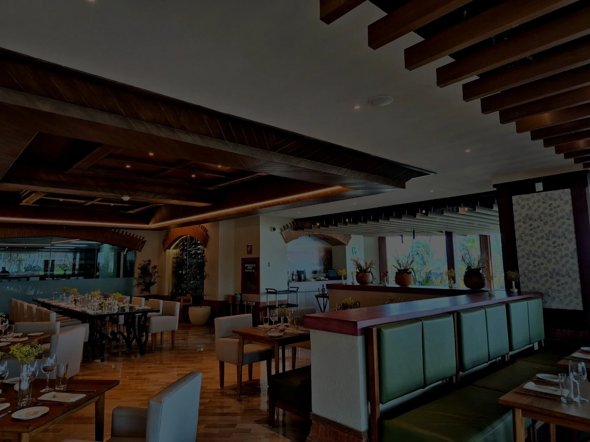 Interior de un restaurante con mesas decoradas con copas y platos, paredes blancas, ventanas grandes y techo con vigas de madera.