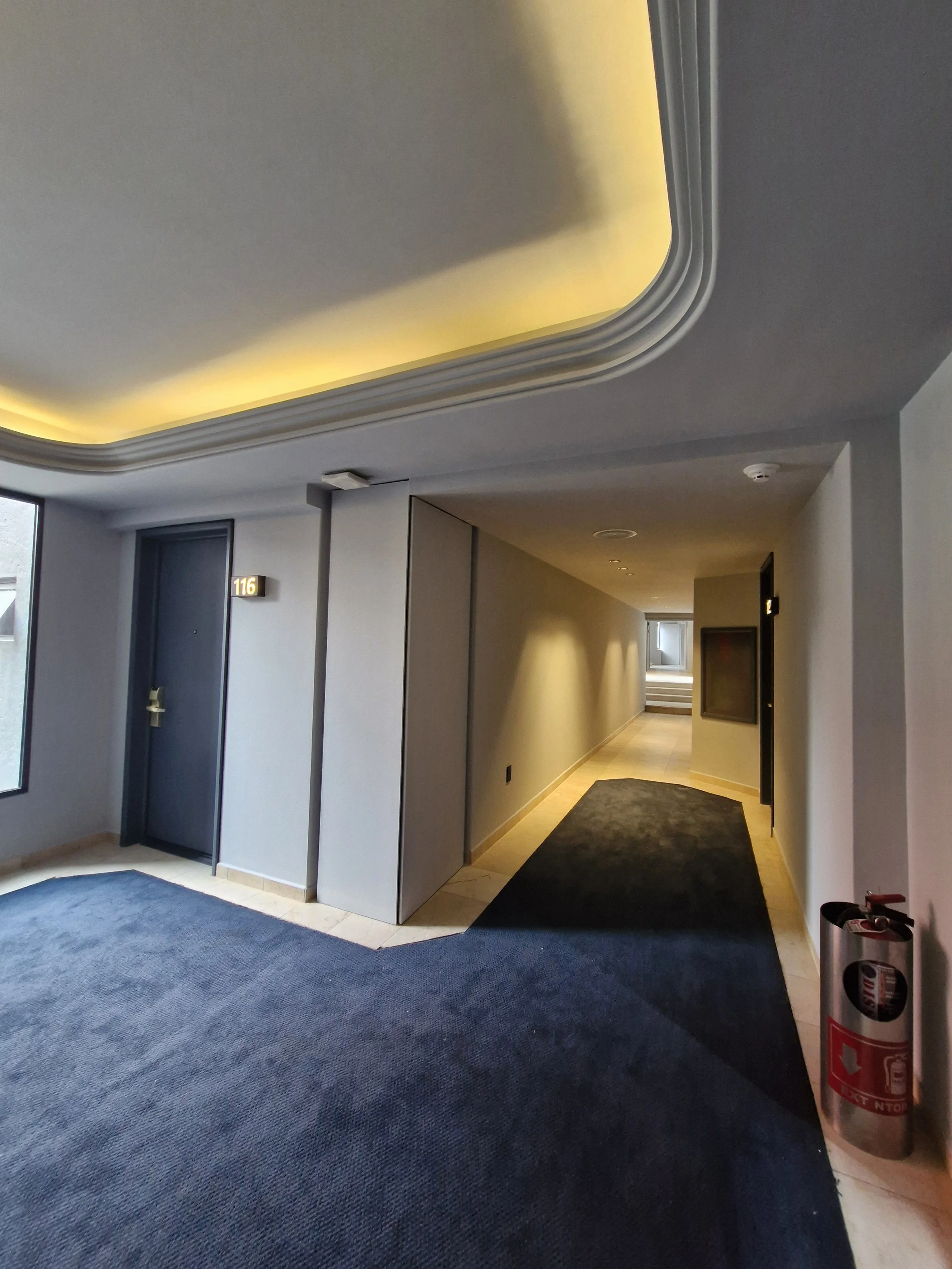 Pasillo de un hotel con puerta número 116, alfombra negra y luz cálida en el techo.