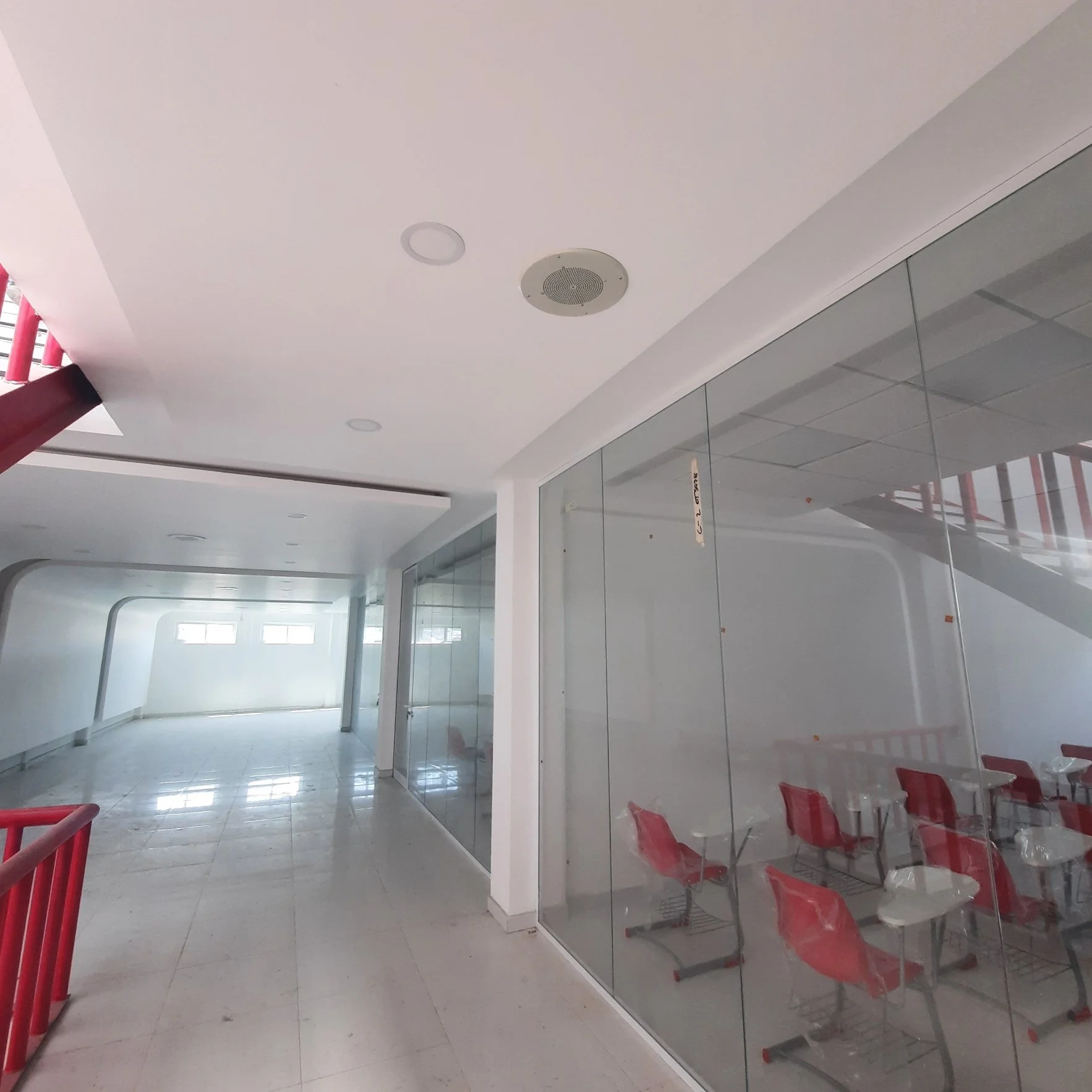 Interior de un espacio comercial con paredes de vidrio y asientos rojos, vista de techo con focos y altavoces, paredes blancas y ventanales que dejan entrar luz natural.