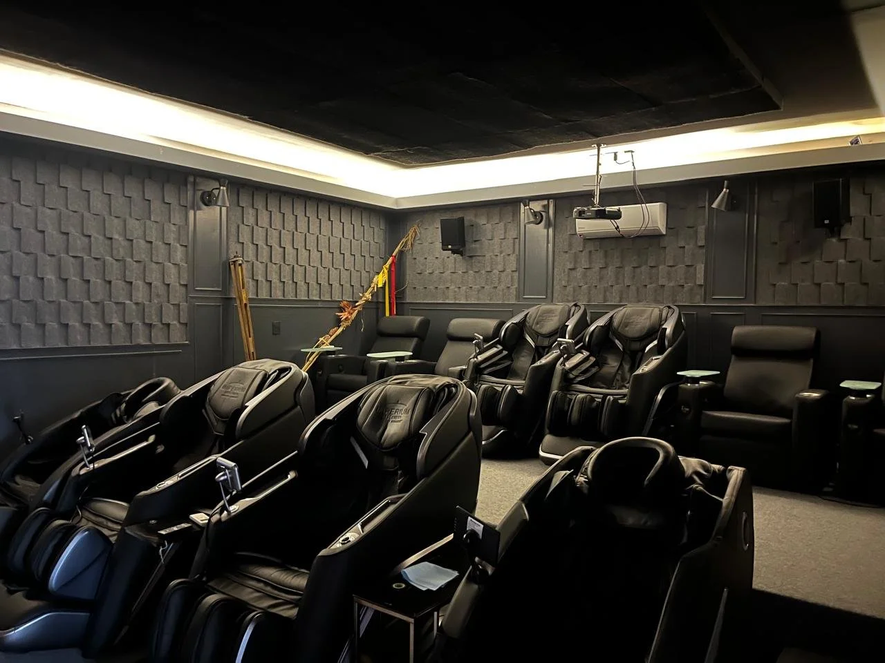 Sala de masajes con sillas de masaje negras y asientos reclinables en una habitación con paredes acústicas grises y techo negro.