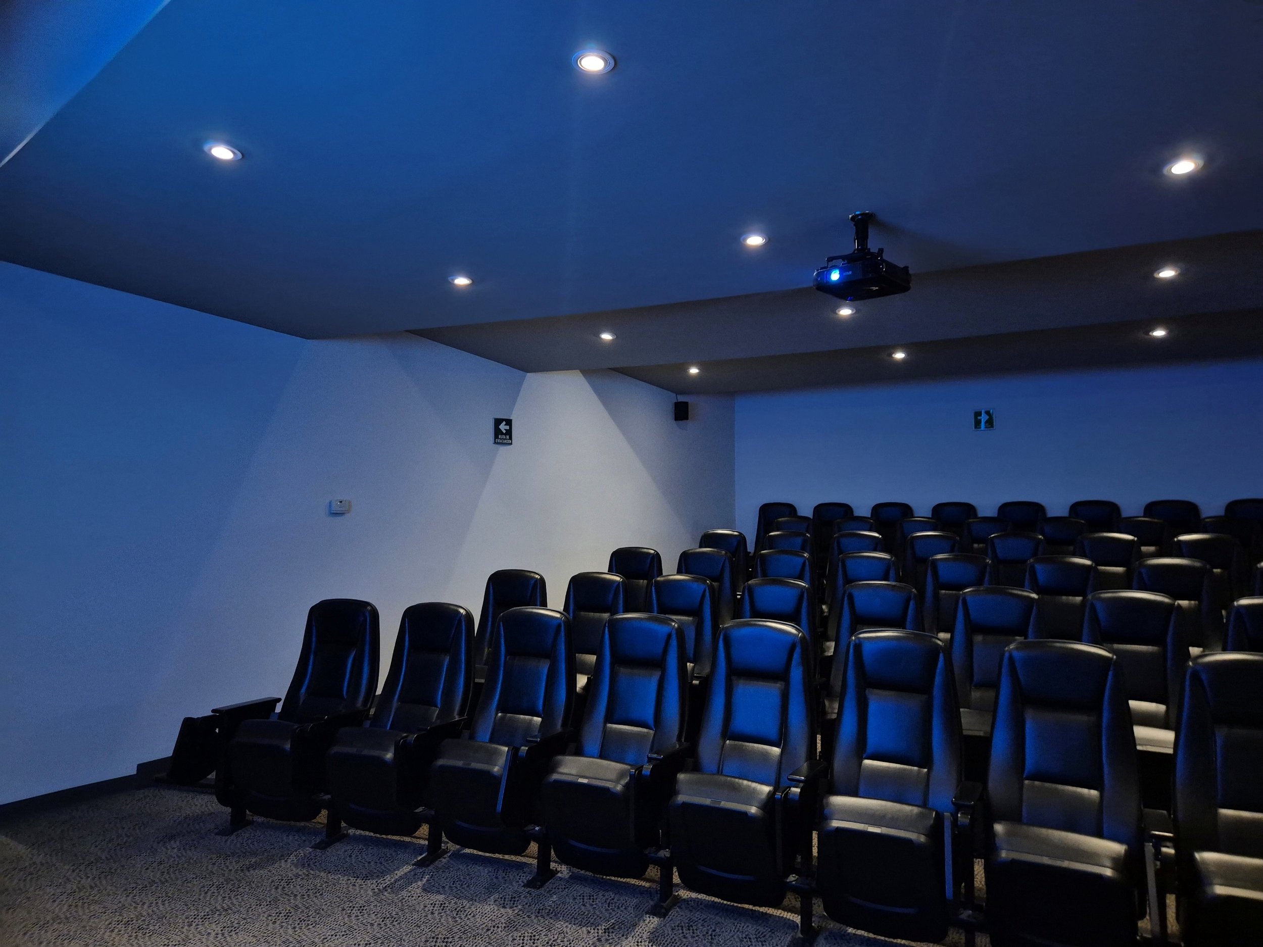 Sala de cine con butacas negras y cortinas azules en las paredes, techo con luces empotradas y proyector en el techo.