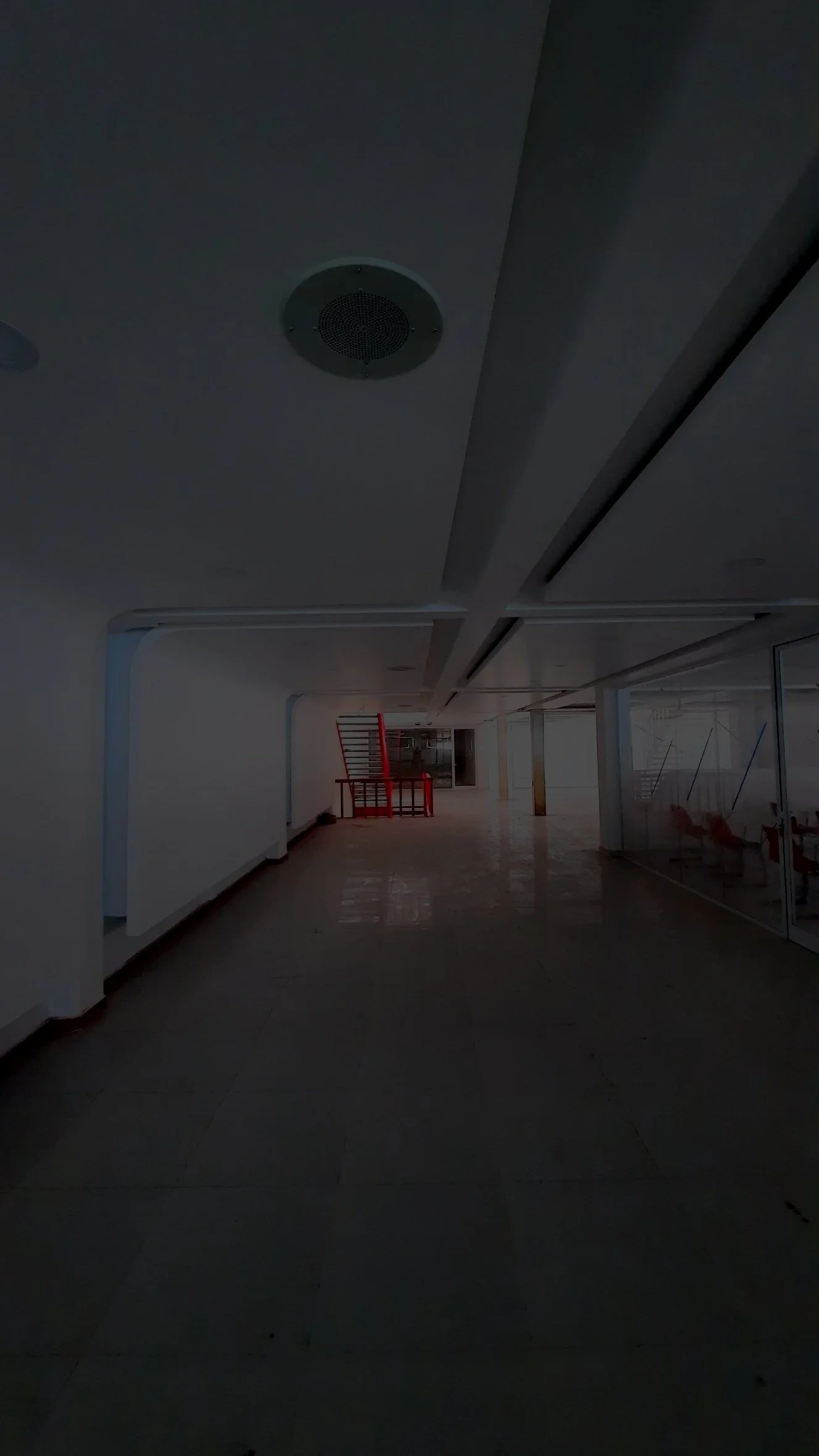 Pasillo vacío en un edificio moderno con paredes blancas, suelo de baldosas y estructura metálica, con una escultura roja en el fondo.