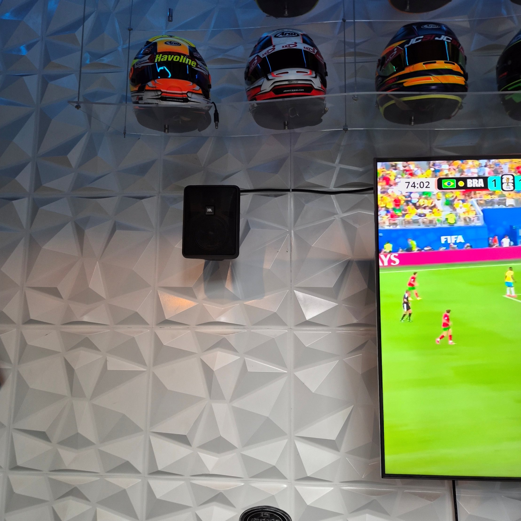 Casco de motocicleta con diseño colorido y letras 'Havoline' en la parte superior, exhibido en un muro con panel de acrílico y fondo de pared con patrón geométrico, debajo de una pantalla que muestra un partido de fútbol con marcador Brasil 1.
