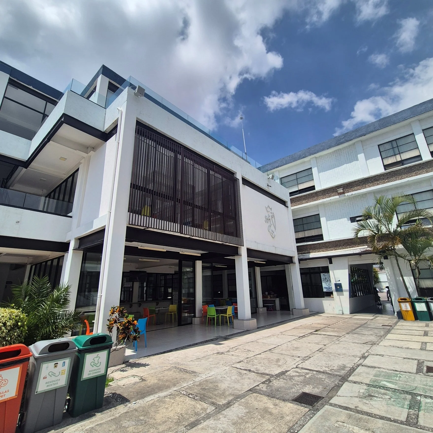 Edificio moderno con área de descanso y mesas en el exterior, árboles decorativos, y colores vibrantes en las sillas. Contenedores de basura para diferentes tipos de residuos en primer plano. Cielo parcialmente nublado.