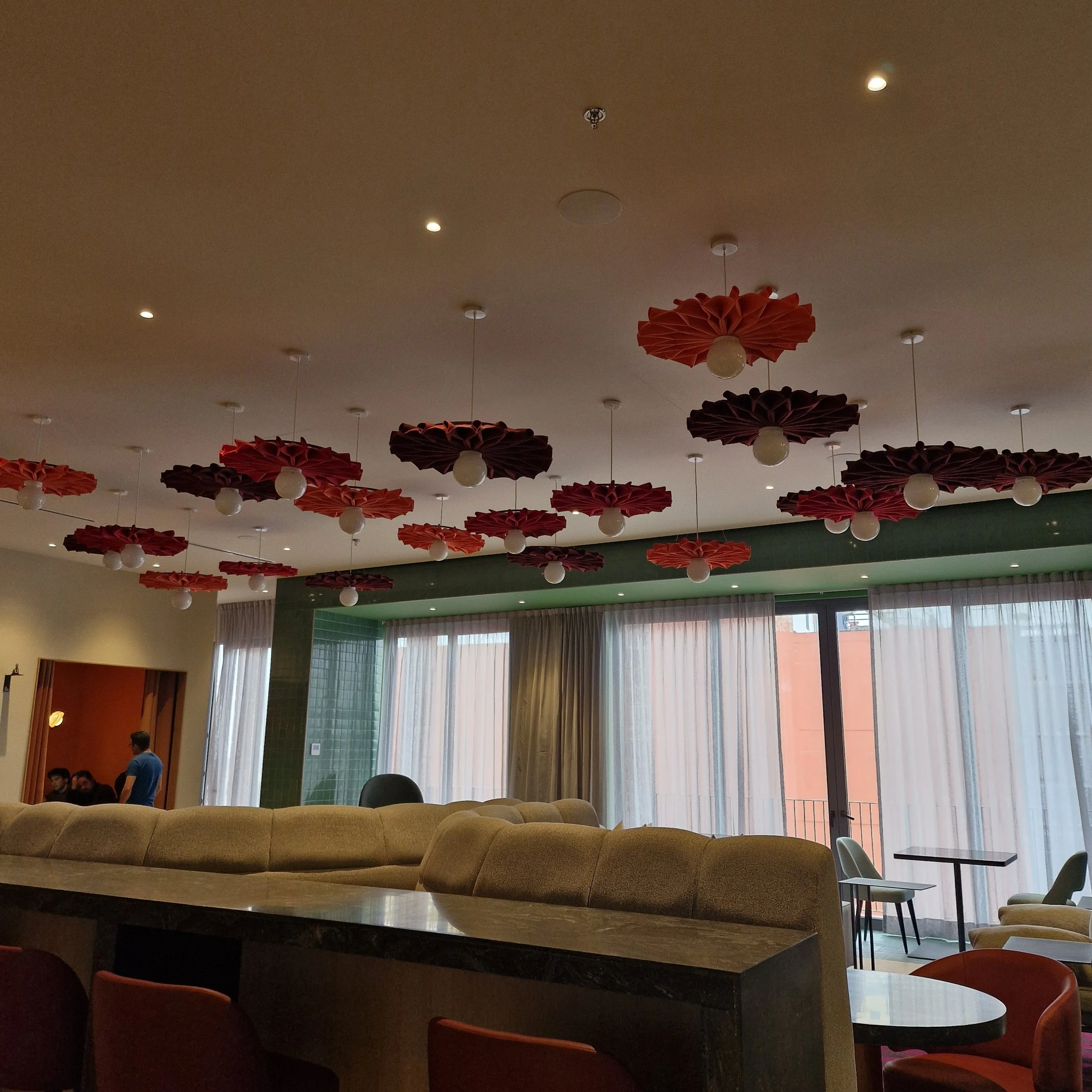 Interior de un espacio moderno con sillones beige, mesas redondas y sillas de colores azul y naranja, con grandes ventanas con cortinas blancas y verdes, decorado con lámparas con pantallas rojas en forma de flor suspendidas del techo