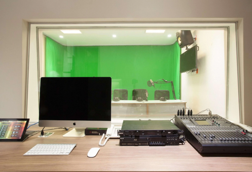 Estudio de grabación con equipo de audio y una computadora, vista desde la cabina con ventana hacia la sala con micrófono, sillas y fondo verde.