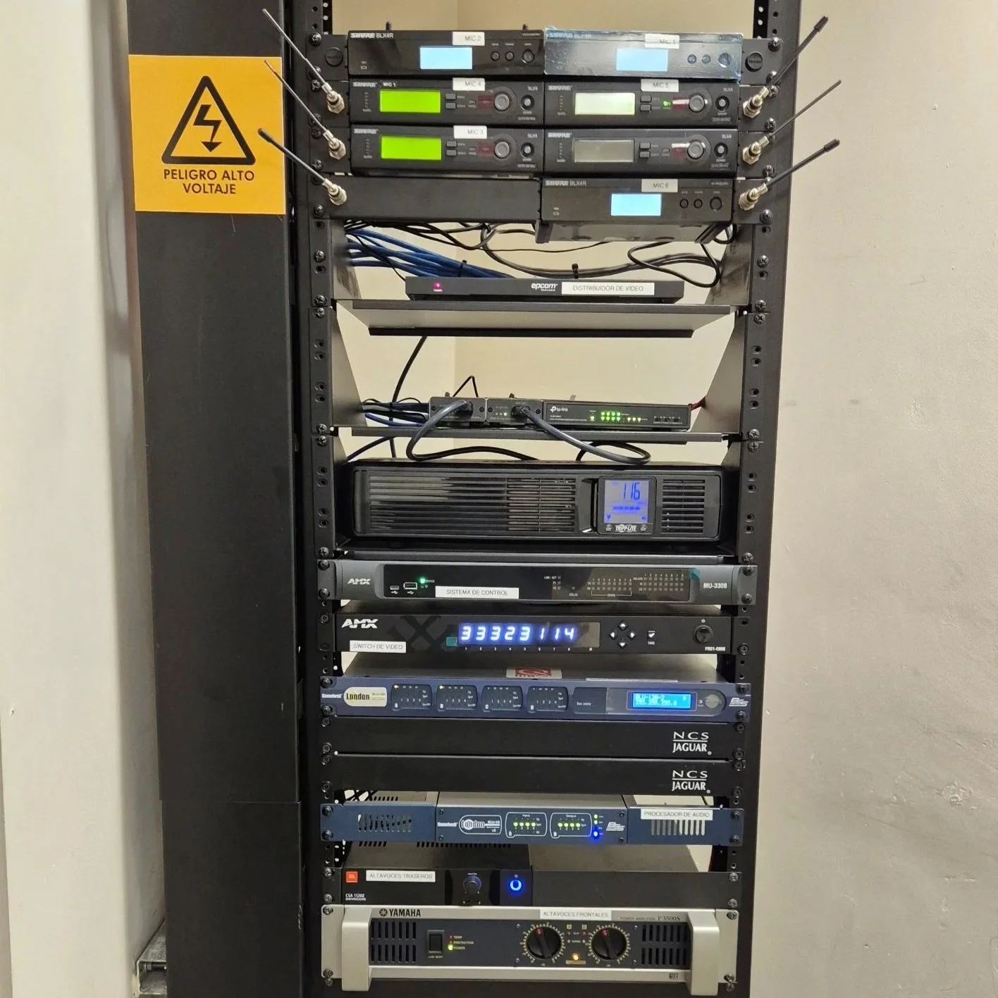 Un equipo de sonido y video en un rack, con múltiples componentes electrónicos y un letrero que dice 'Peligro Alto Voltaje'.