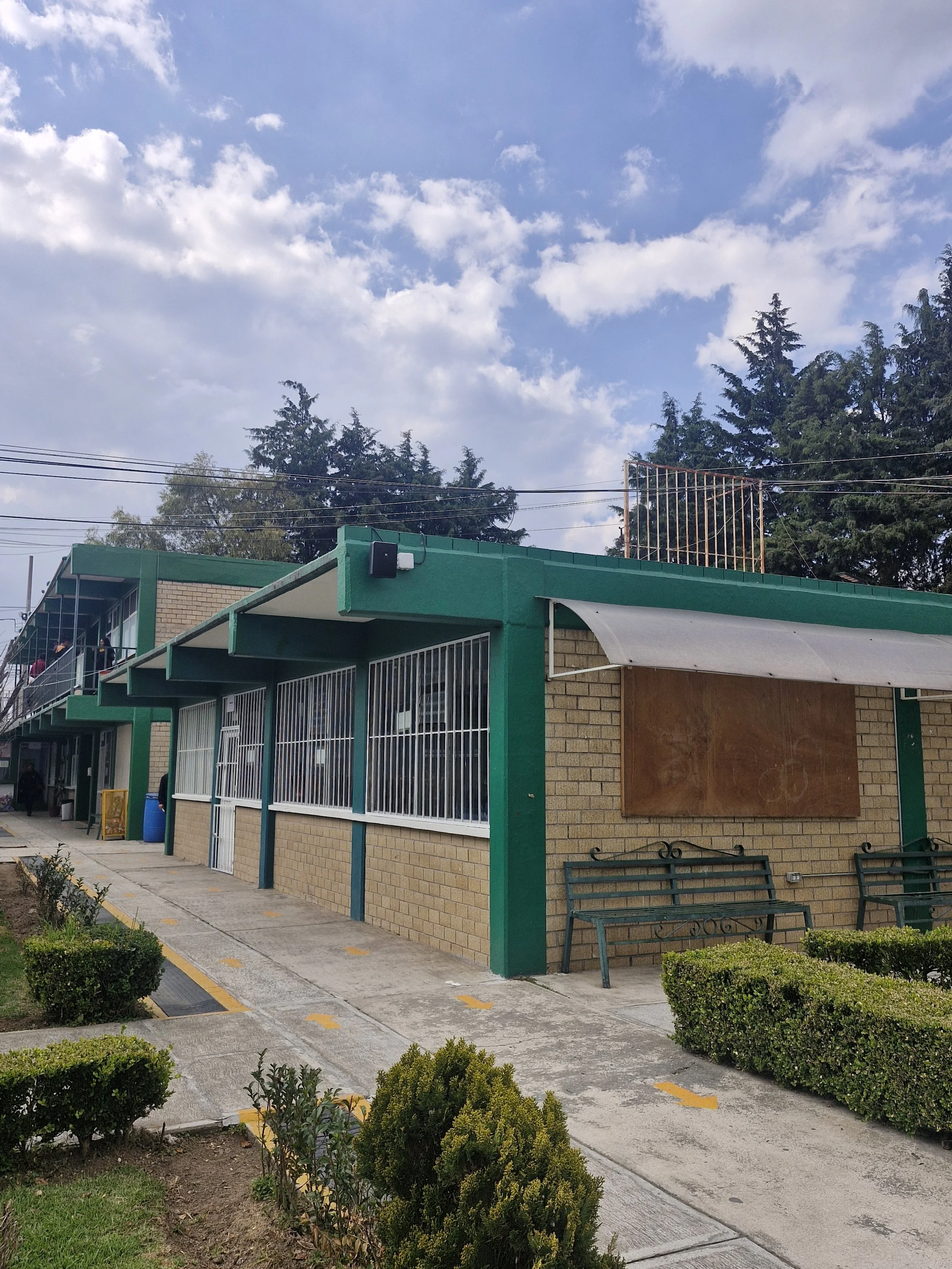 Edificio con paredes de ladrillo y estructura de metal verde, ventanas con rejas, un toldo blanco y un cartel vacío en una acera con arbustos y un banco cerca.