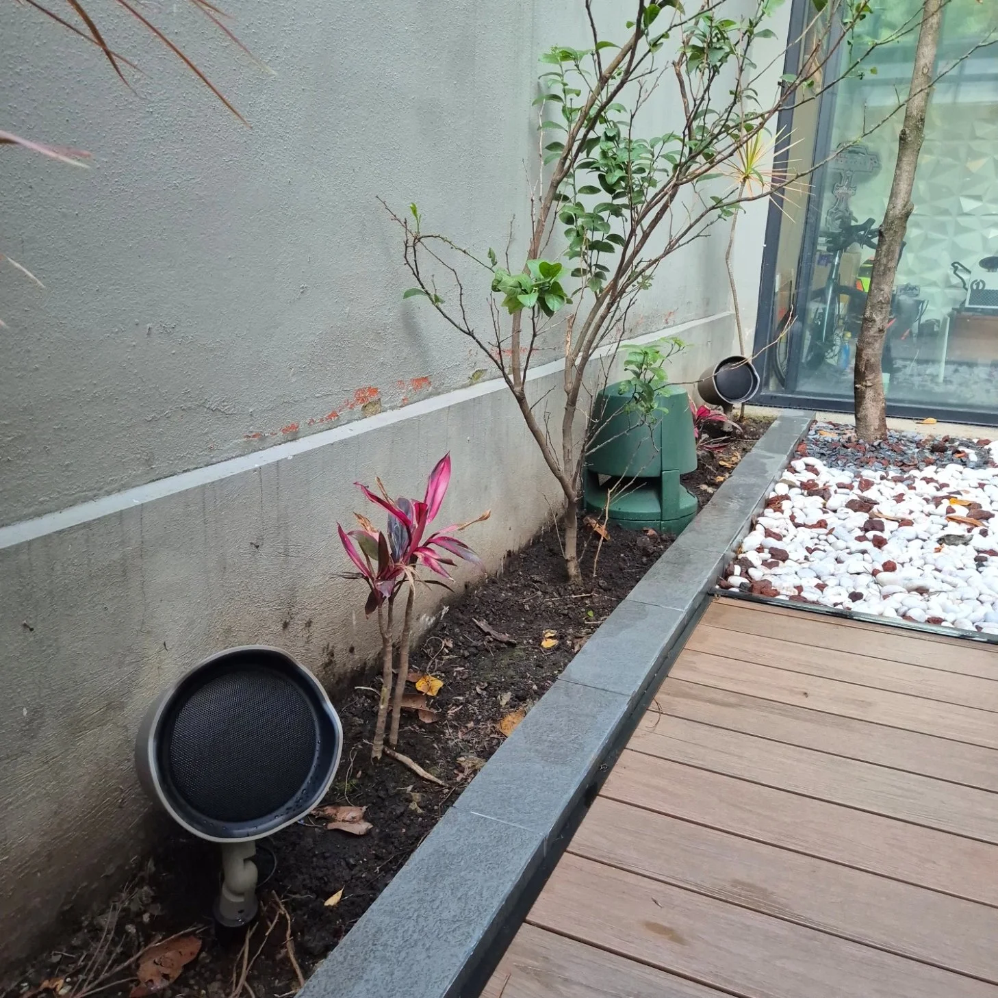 Jardín con plantas y flores, con área de piedra blanca, madera y pared gris, en un espacio exterior de casa.