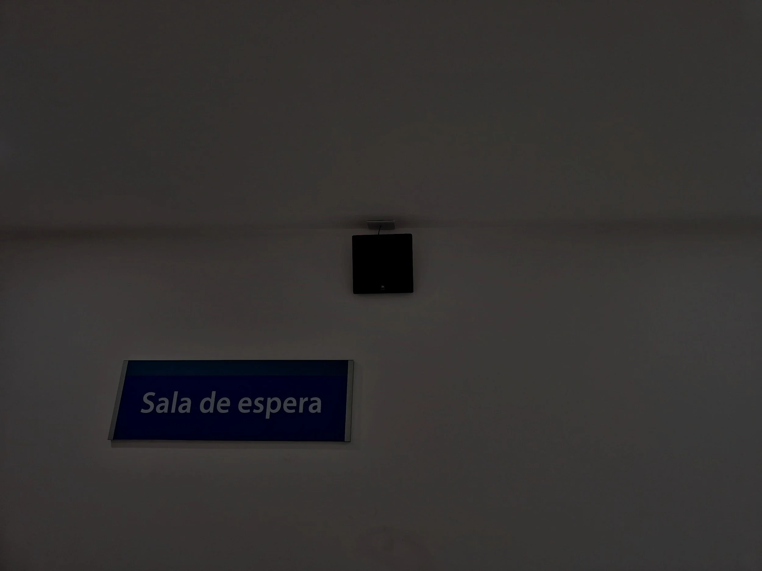 Imagen de un cartel que dice 'Sala de espera' colgado en una pared, junto a un monitor y un aparato de sonido en una sala interior