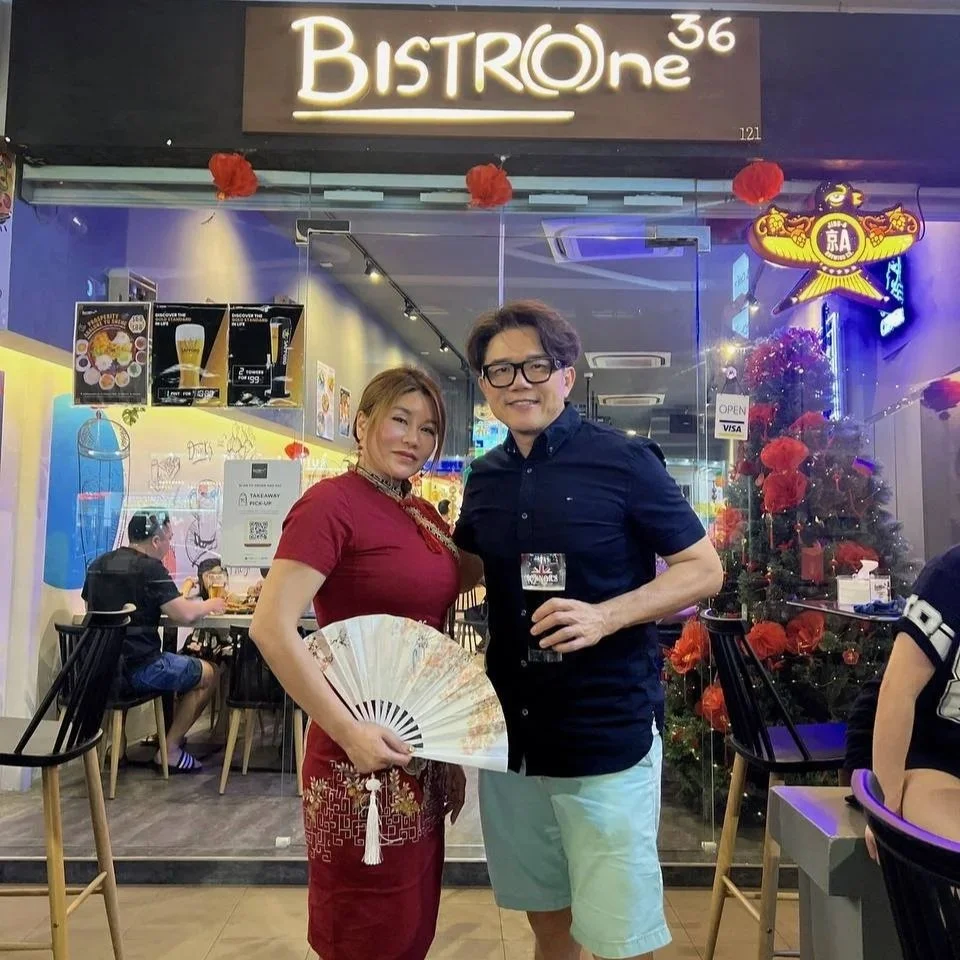 A woman and a man stand inside a restaurant, smiling at the camera. The woman is dressed in a red dress and is holding a decorative fan. The man is wearing glasses and a dark shirt and is holding a glass of drink. The restaurant sign above reads "BIS