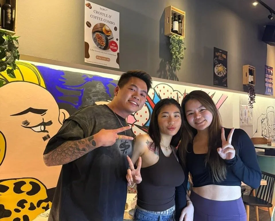 Three friends smiling and making peace signs in a colorful cafe with cartoon-style wall art and posters.