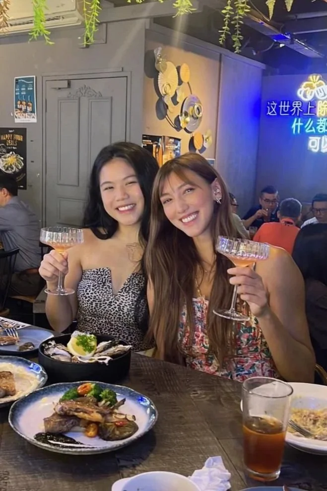Two young women smiling and holding cocktail glasses at a restaurant table with food and drinks, with a dimly lit restaurant interior in the background.