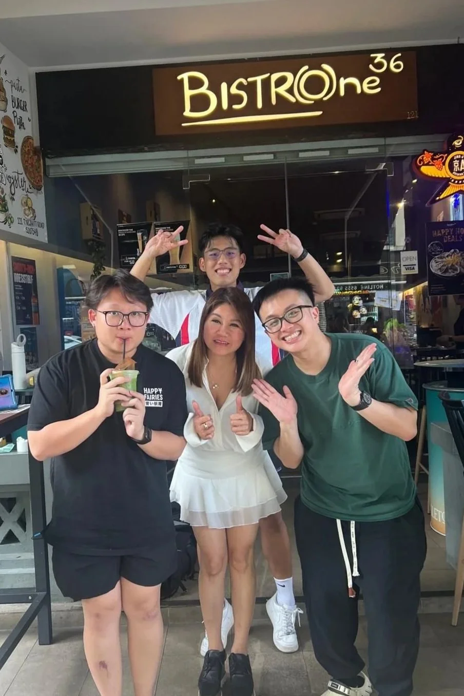 A group of four young people posing in front of a restaurant called Bistro One 36, with one holding a drink and others smiling and making gestures inside a casual dining environment. Getfed, overkill