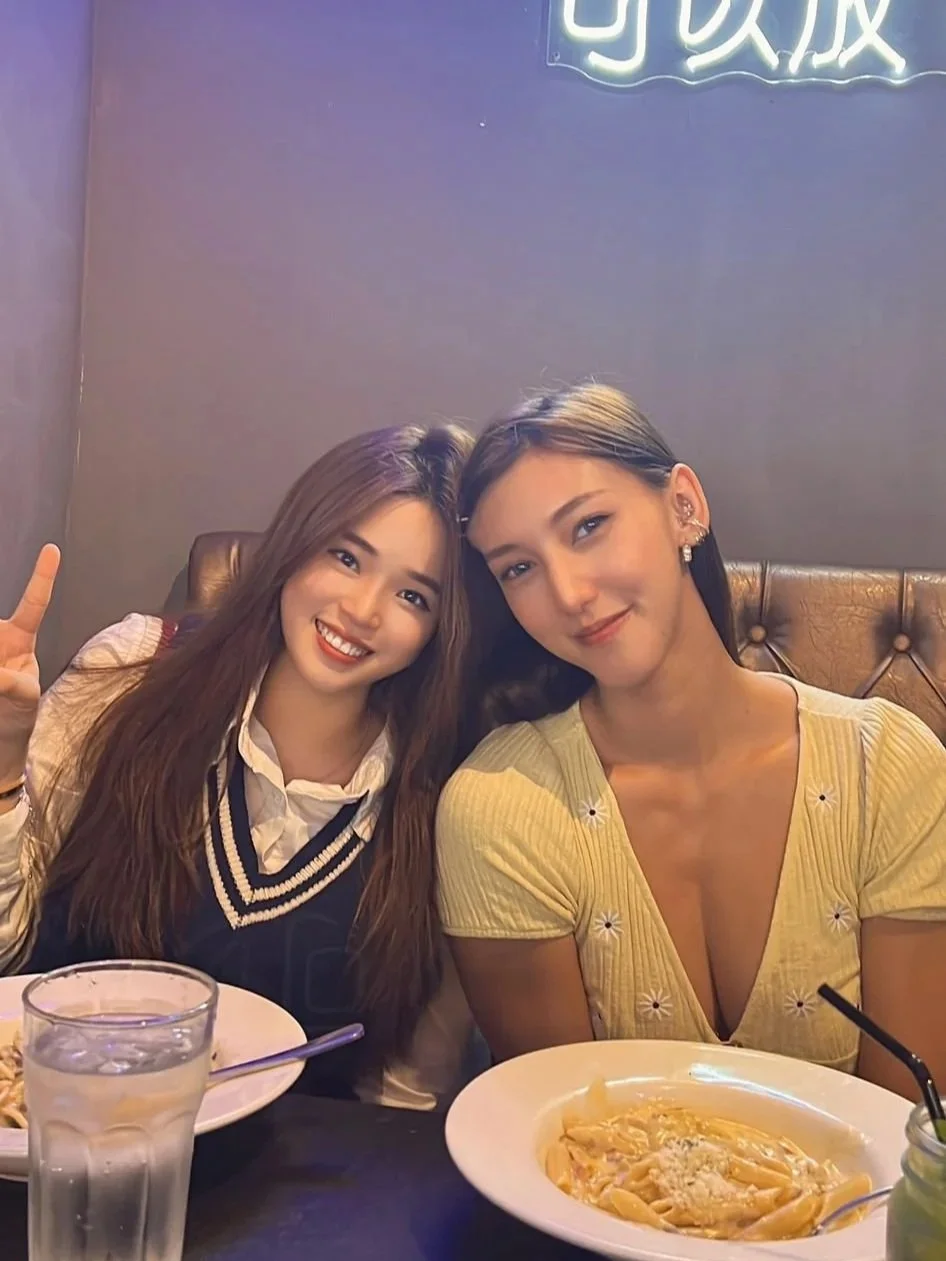 Two young women sitting at a restaurant table with plates of pasta, smiling at the camera, one making a peace sign, illuminated by neon sign lighting