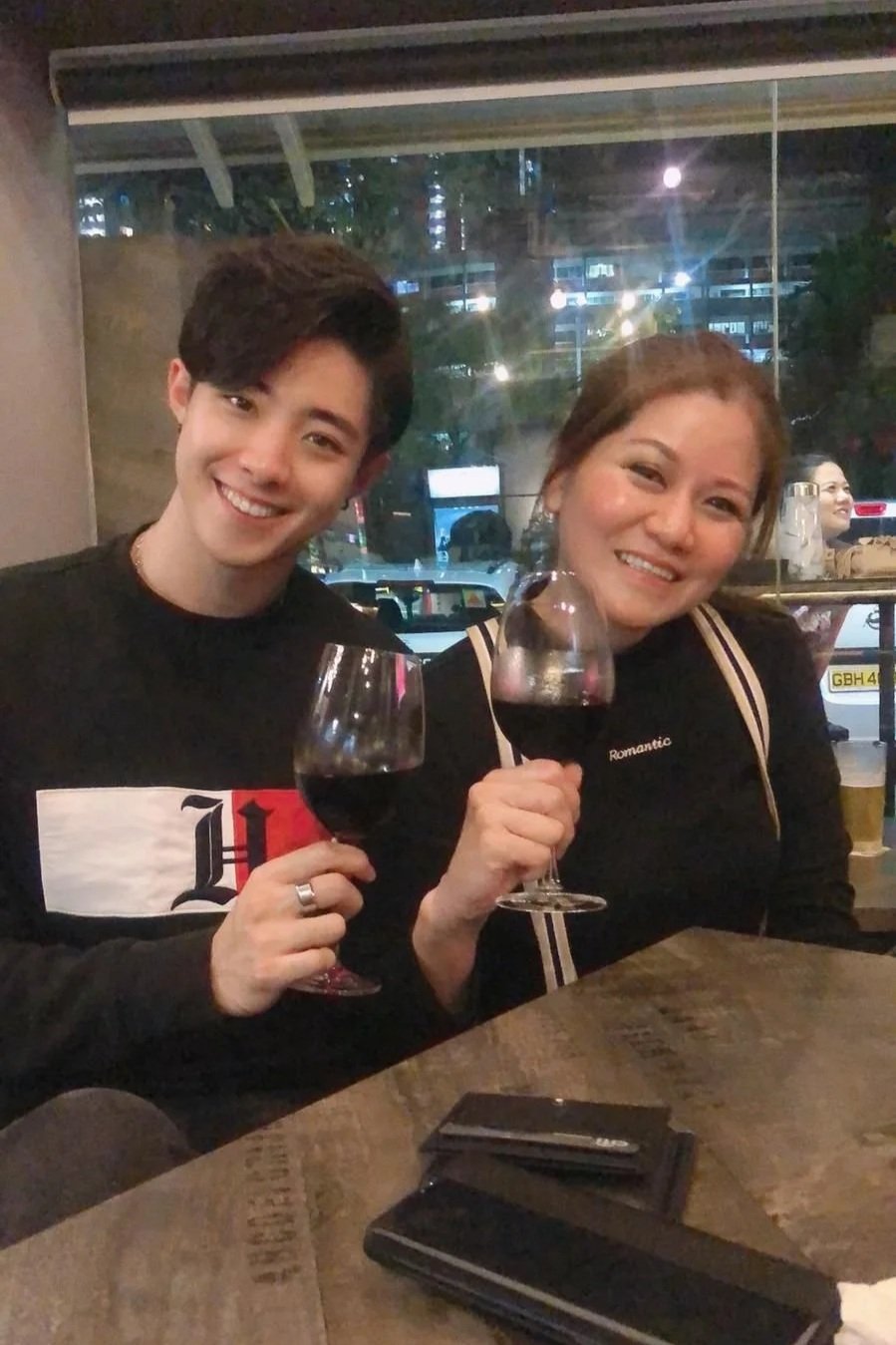 A young man and a woman sit at a table in a restaurant, smiling and holding glasses of red wine. The woman is wearing a black shirt with the word 'Romantic' on it. The background shows large windows with a city street and some people outside.