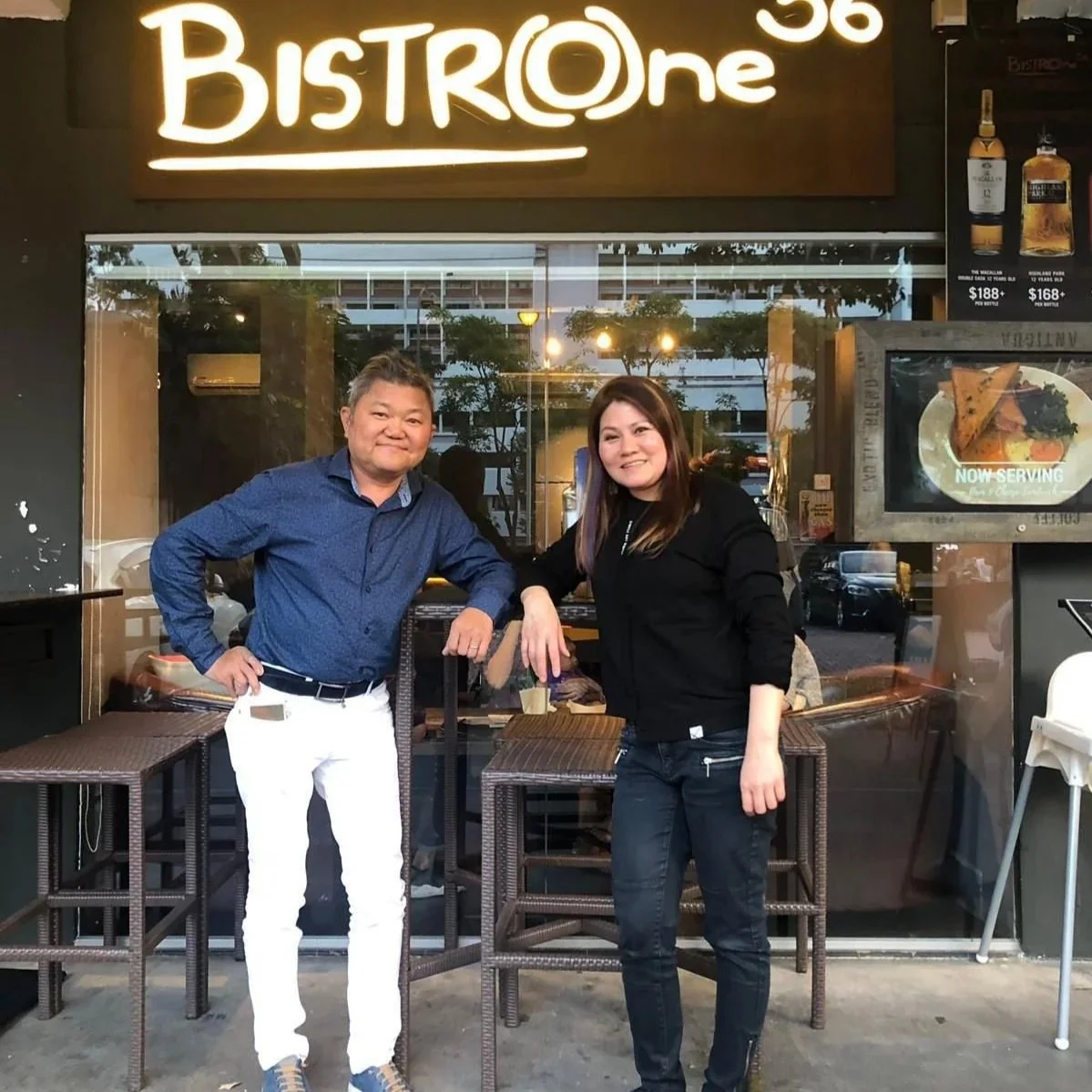Two people standing outside a restaurant called Bistro One, smiling at the camera. The person on the left is wearing a blue shirt and white pants, while the person on the right is wearing black clothing. The restaurant has a sign that says 'NOW SERVI