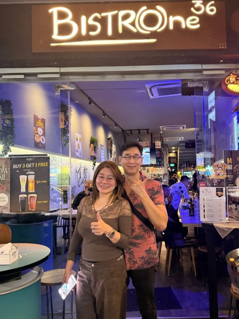 Two smiling people, a woman and a man, standing in front of a restaurant named BISTROne. The woman is giving a thumbs up while the man is touching his chin. Inside the restaurant, there are people dining and various signs and menus visible.
