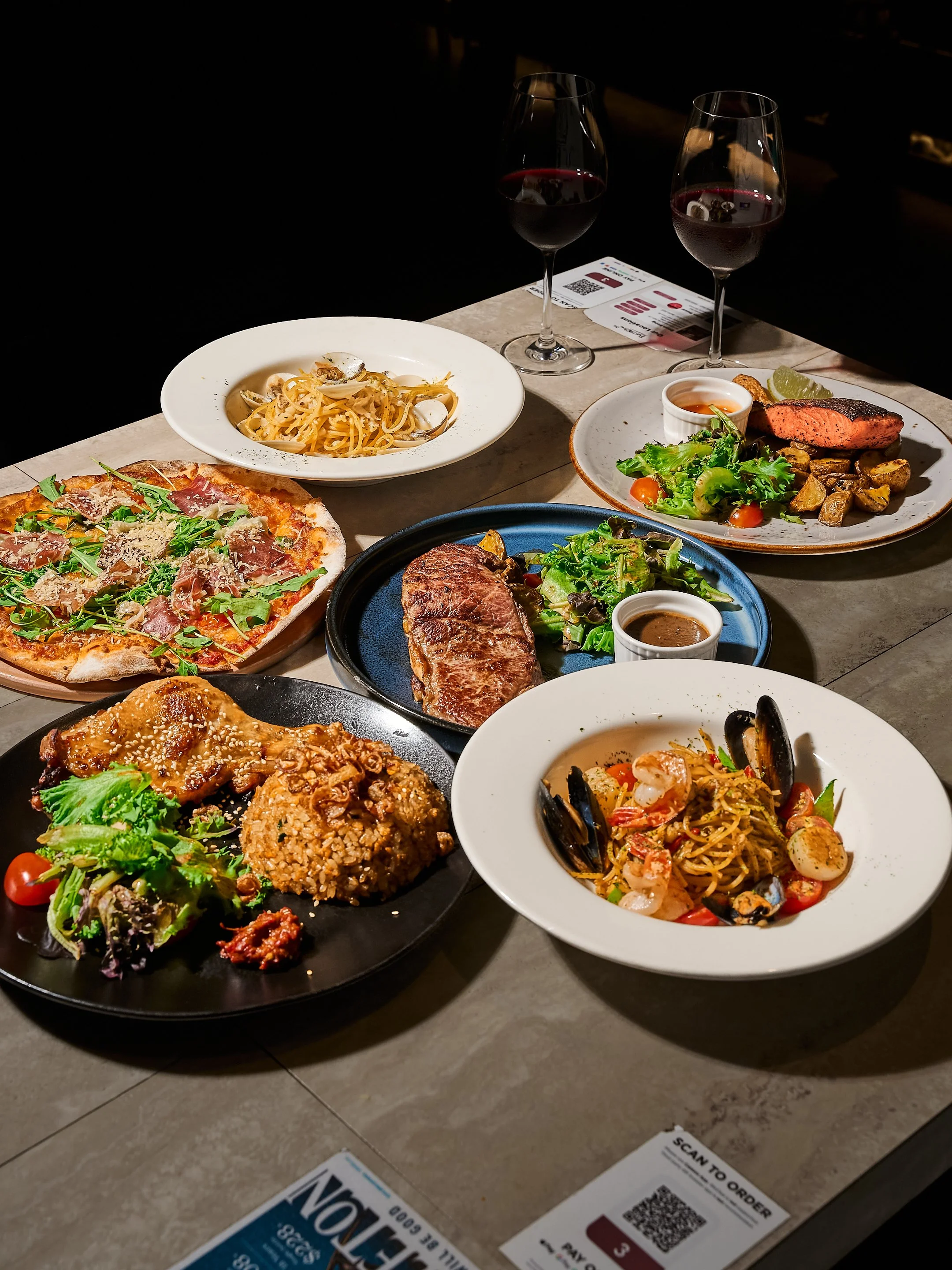 A table with various dishes of food and two glasses of red wine at a restaurant.