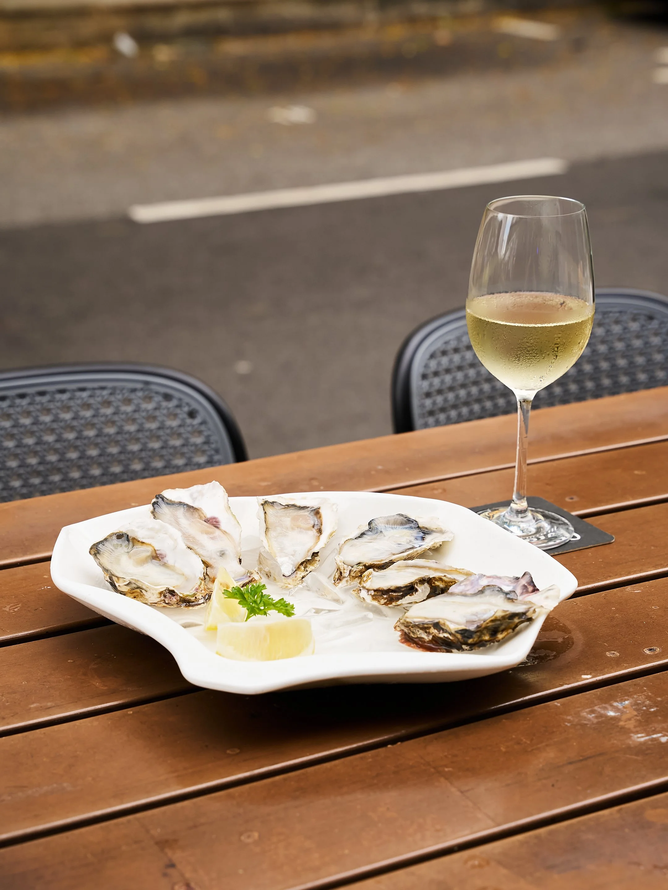 A plate of oysters with lemon wedges and parsley, a glass of white wine on a wooden table outdoors.