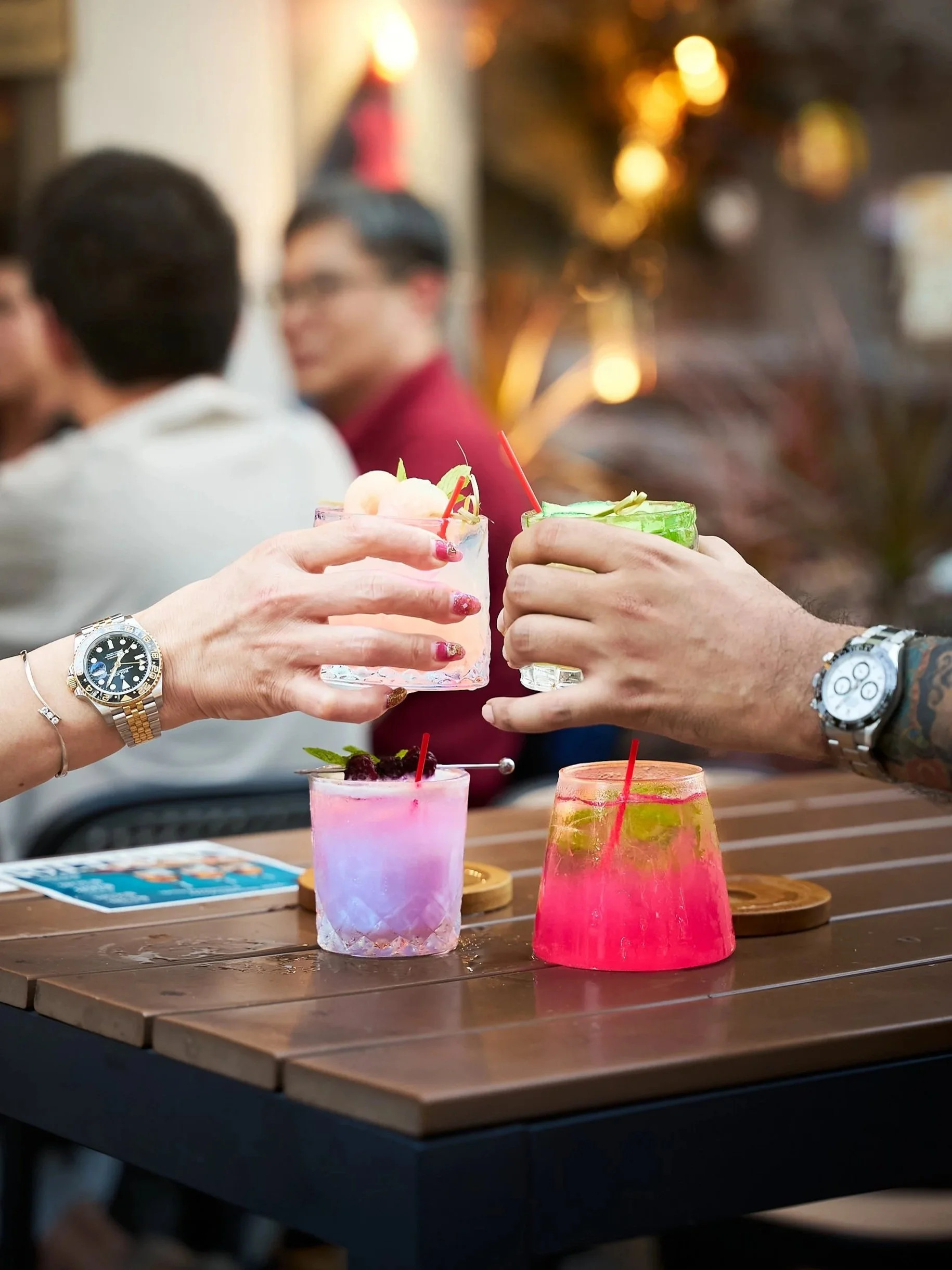 People clinking colorful drinks at a bar or restaurant, with a blurred background of warm lights and other patrons.