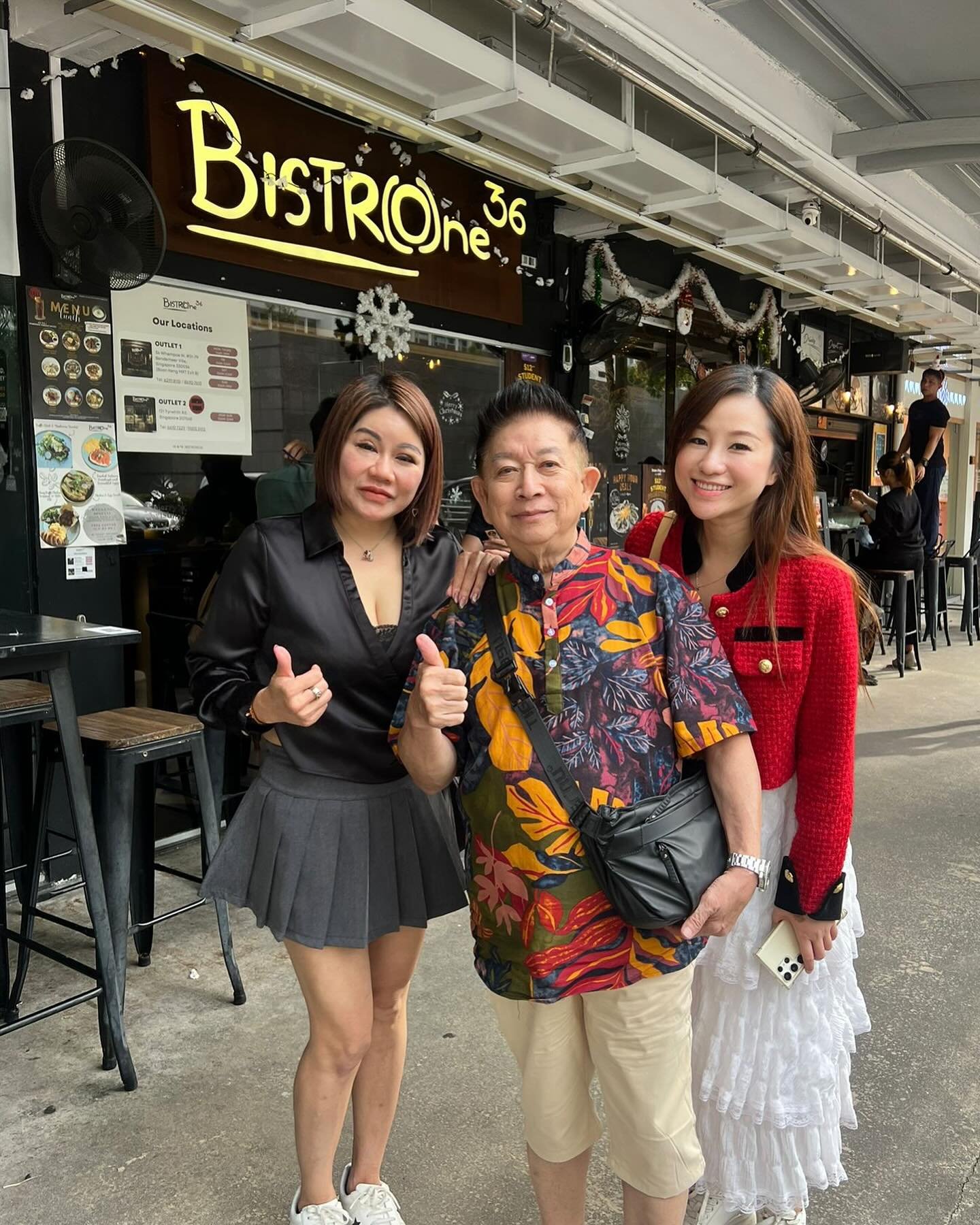 Three people standing outside a restaurant named Bistrone, smiling and posing for the photo. The restaurant's sign is visible behind them, decorated with Christmas ornaments.