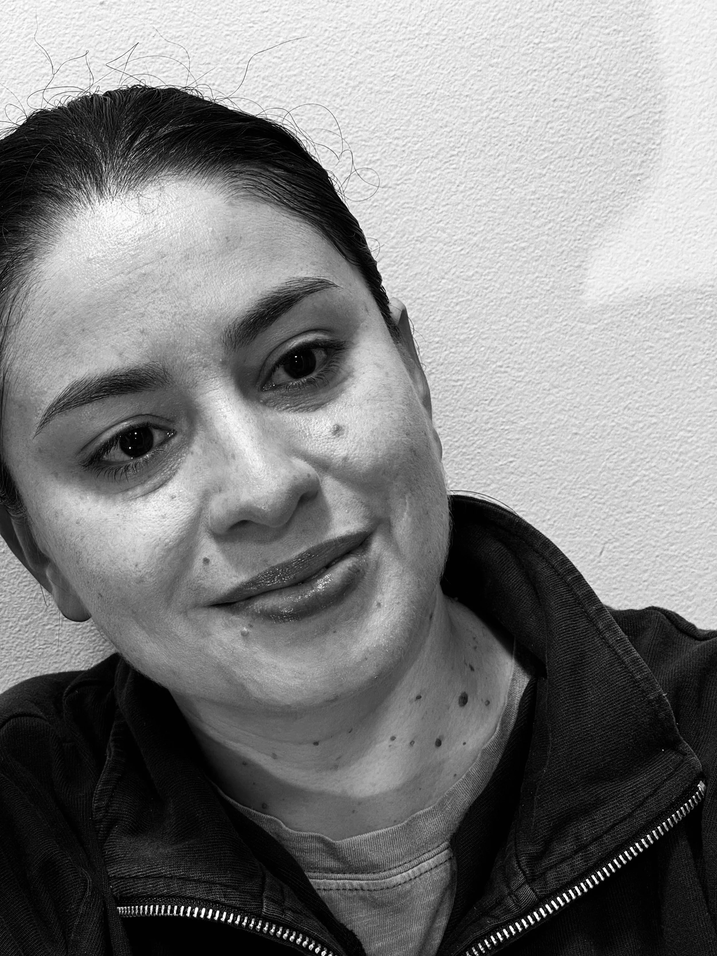 Close-up black and white photo of a woman with dark hair, smiling, wearing a zip-up jacket and a striped shirt, against a plain wall.