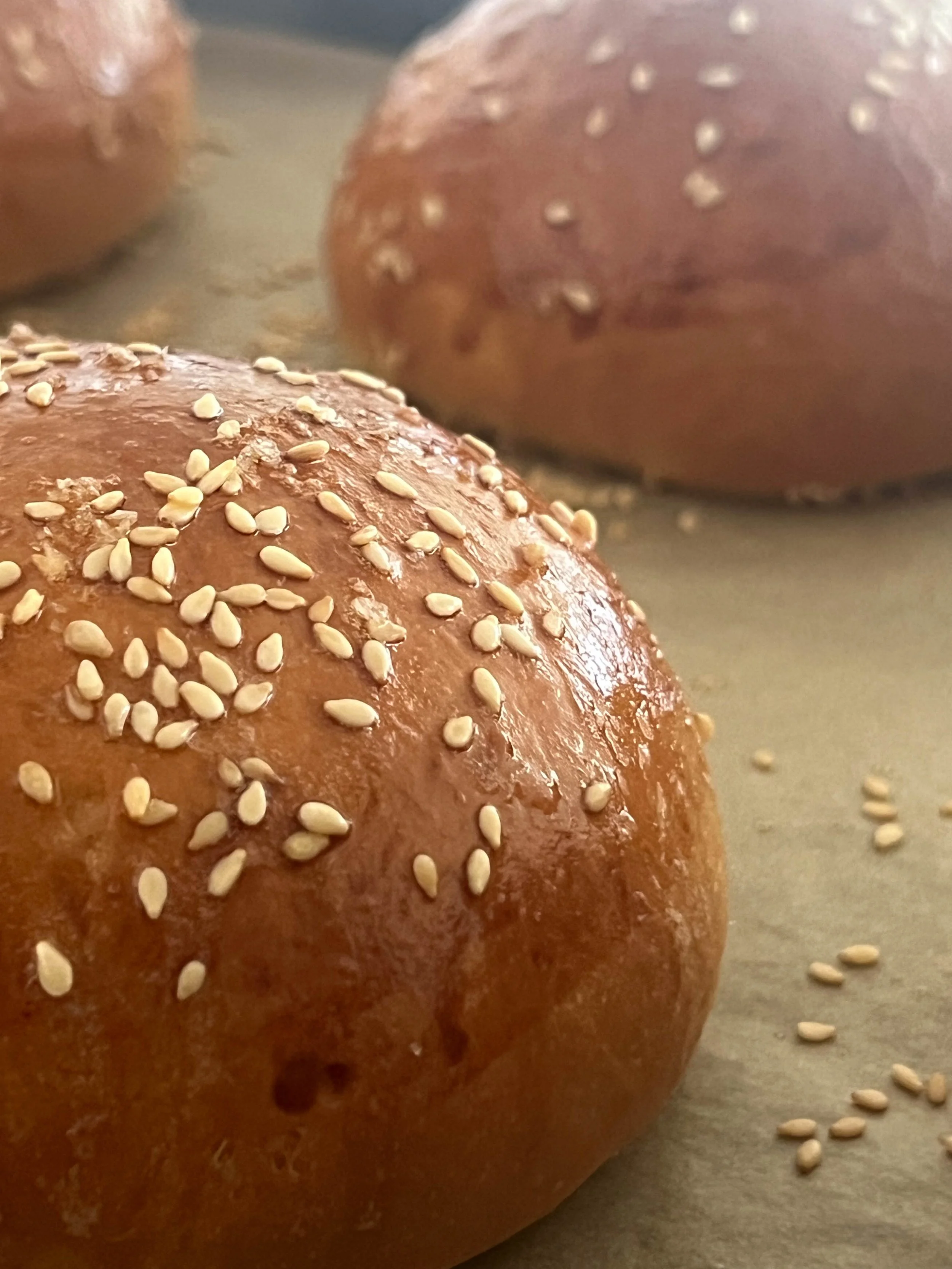 Close-up of a sesame seed bun with sesame seeds on top, with additional sesame seeds scattered on a flat surface nearby.