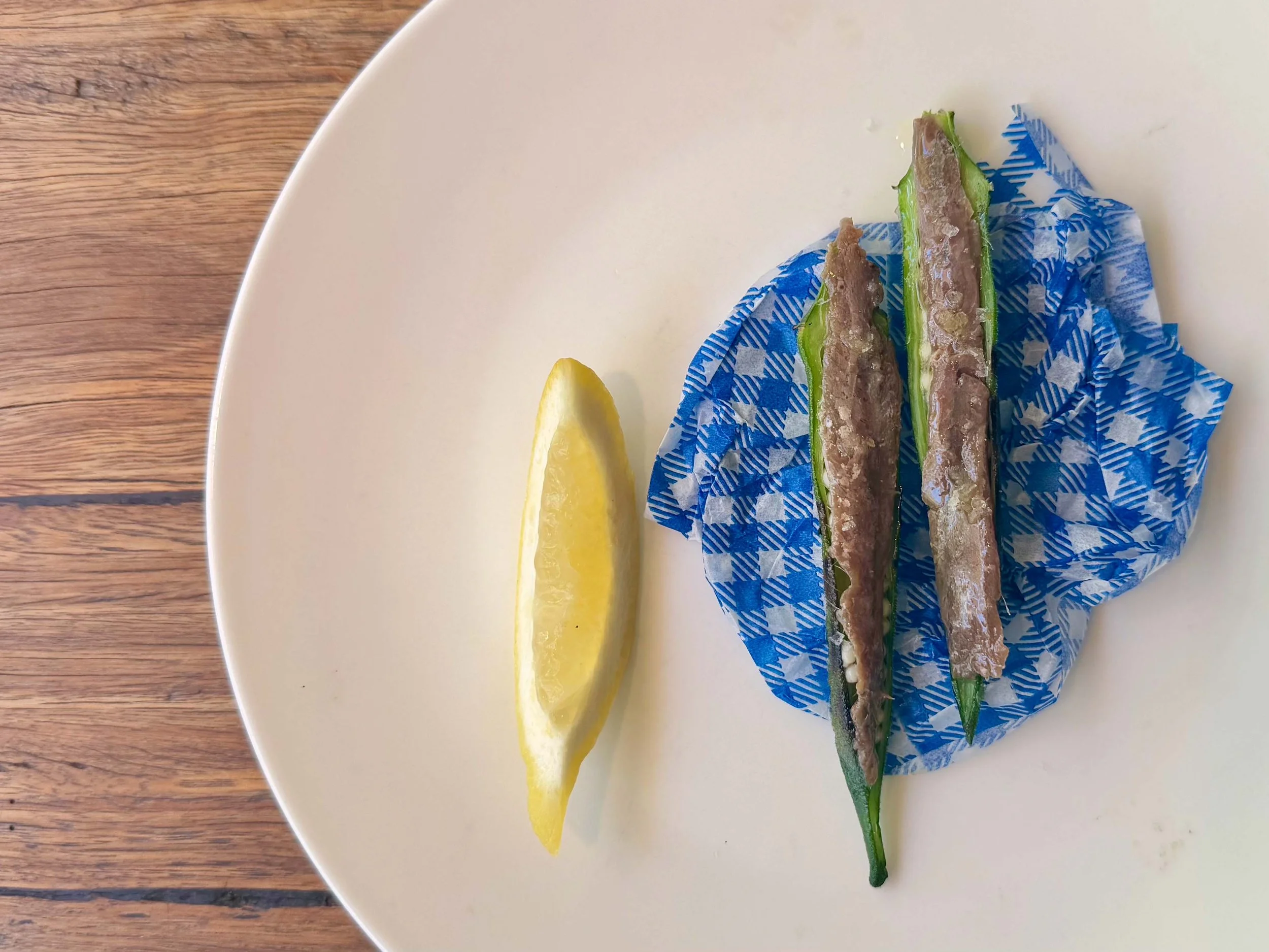 A white plate contains three slices of cucumber topped with a grayish pate-like spread, placed on blue and white patterned paper, and a lemon wedge on the side, on a wooden surface.