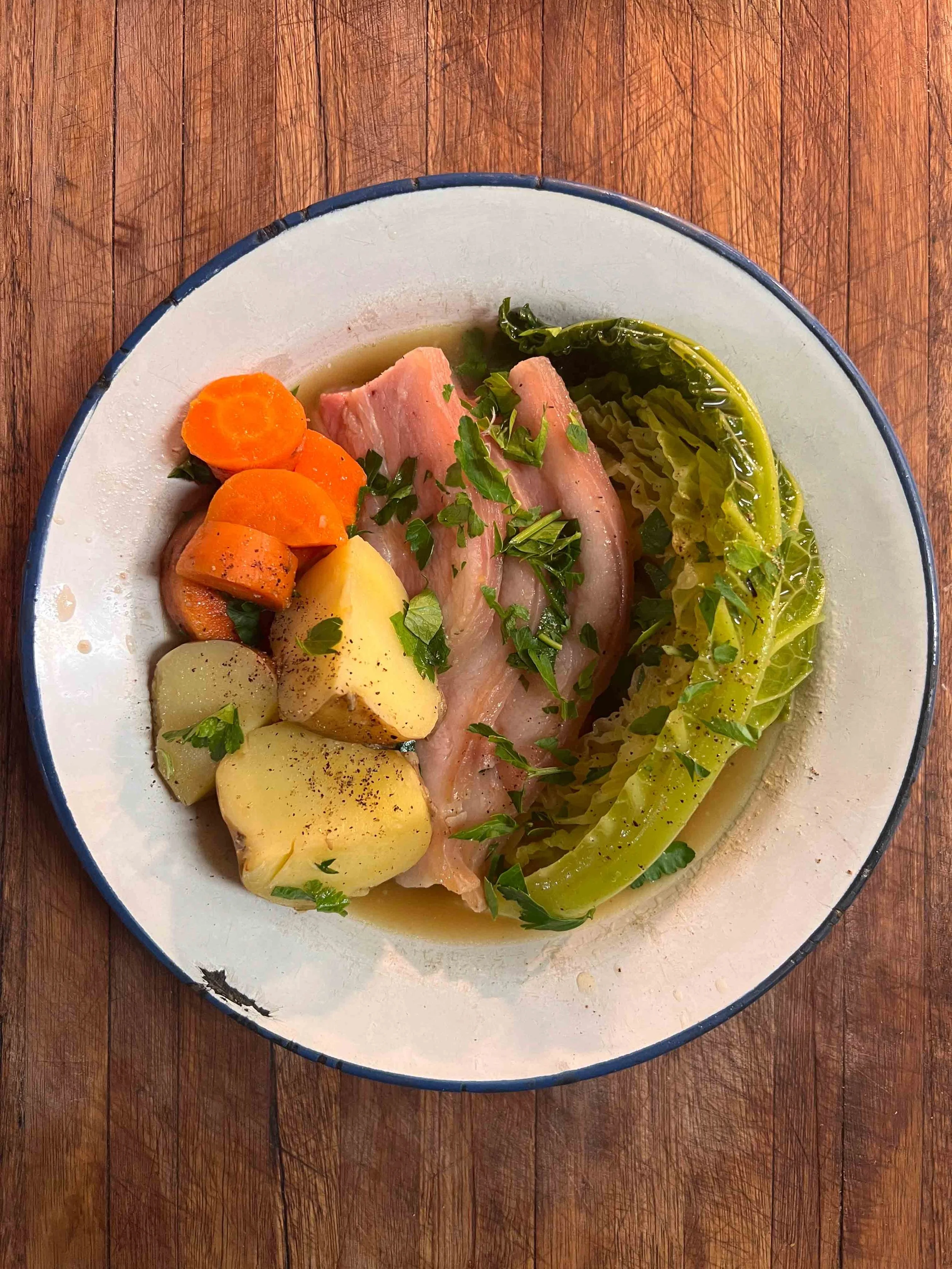 A bowl of hearty chicken soup with carrots, potatoes, leafy greens, and herbs, set on a rustic wooden table.