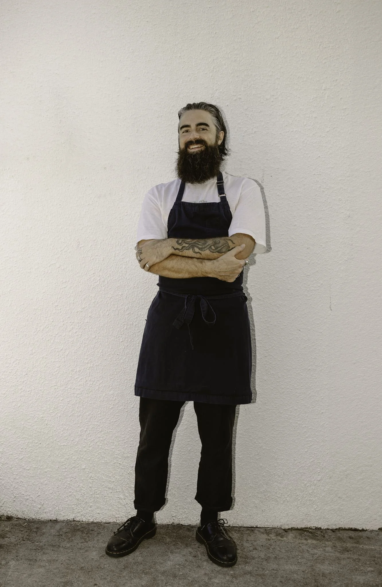 A man with a beard and tattoos on his forearms, wearing a white t-shirt, a black apron, black pants, and black shoes, standing with arms crossed against a plain white wall.