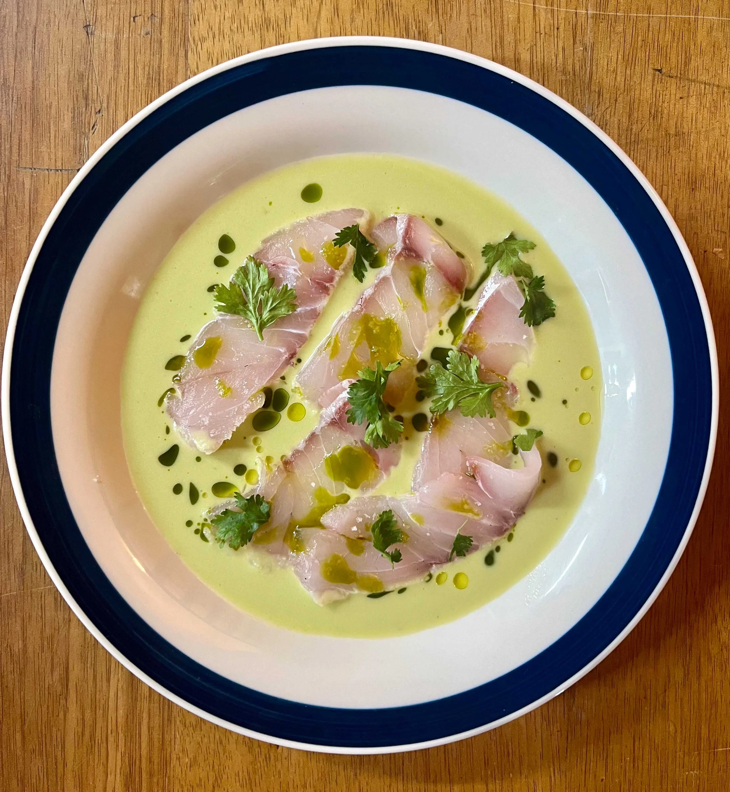 A bowl of green soup topped with slices of raw fish, cilantro leaves, and drizzles of green oil or sauce, served on a wooden surface.