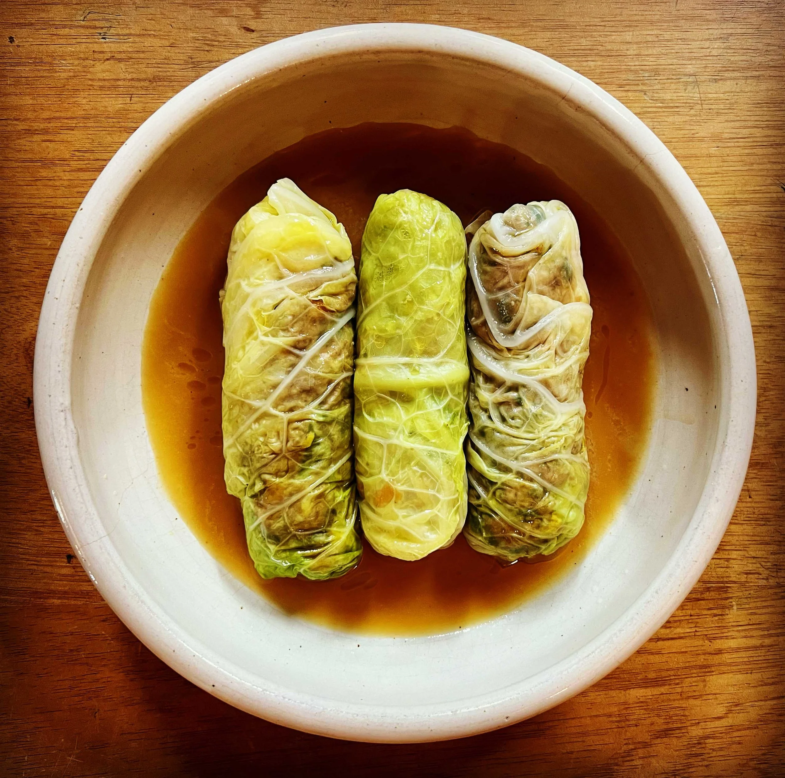 Four stuffed cabbage rolls in a white bowl with sauce on a wooden surface.