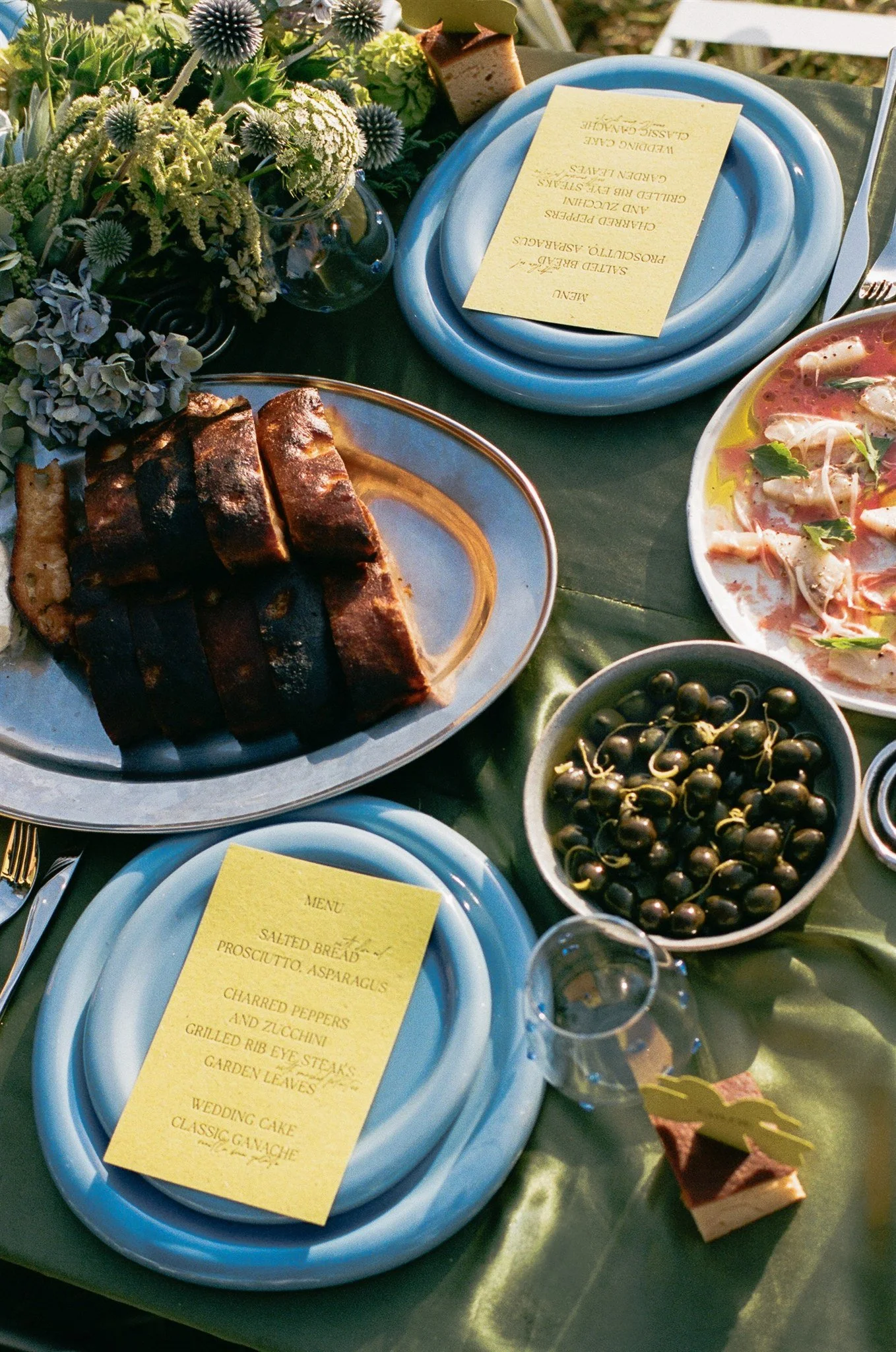 A table setting with a plate of sliced smoked beef brisket, a bowl of black cherries, and a plate of pasta with chicken and vegetables. There is a vase of white and purple flowers, and yellow menu cards on blue plates.