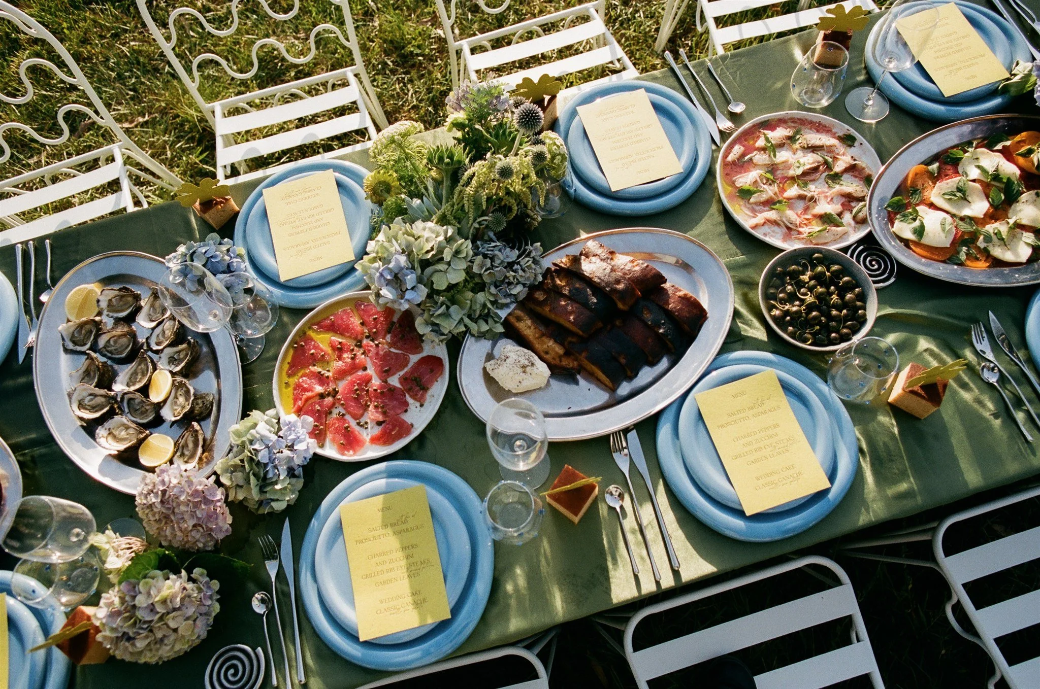 An outdoor dining table set for a meal with blue plates, silverware, wine glasses, and yellow menus, decorated with floral arrangements and appetizers including oysters, prosciutto, and capers.