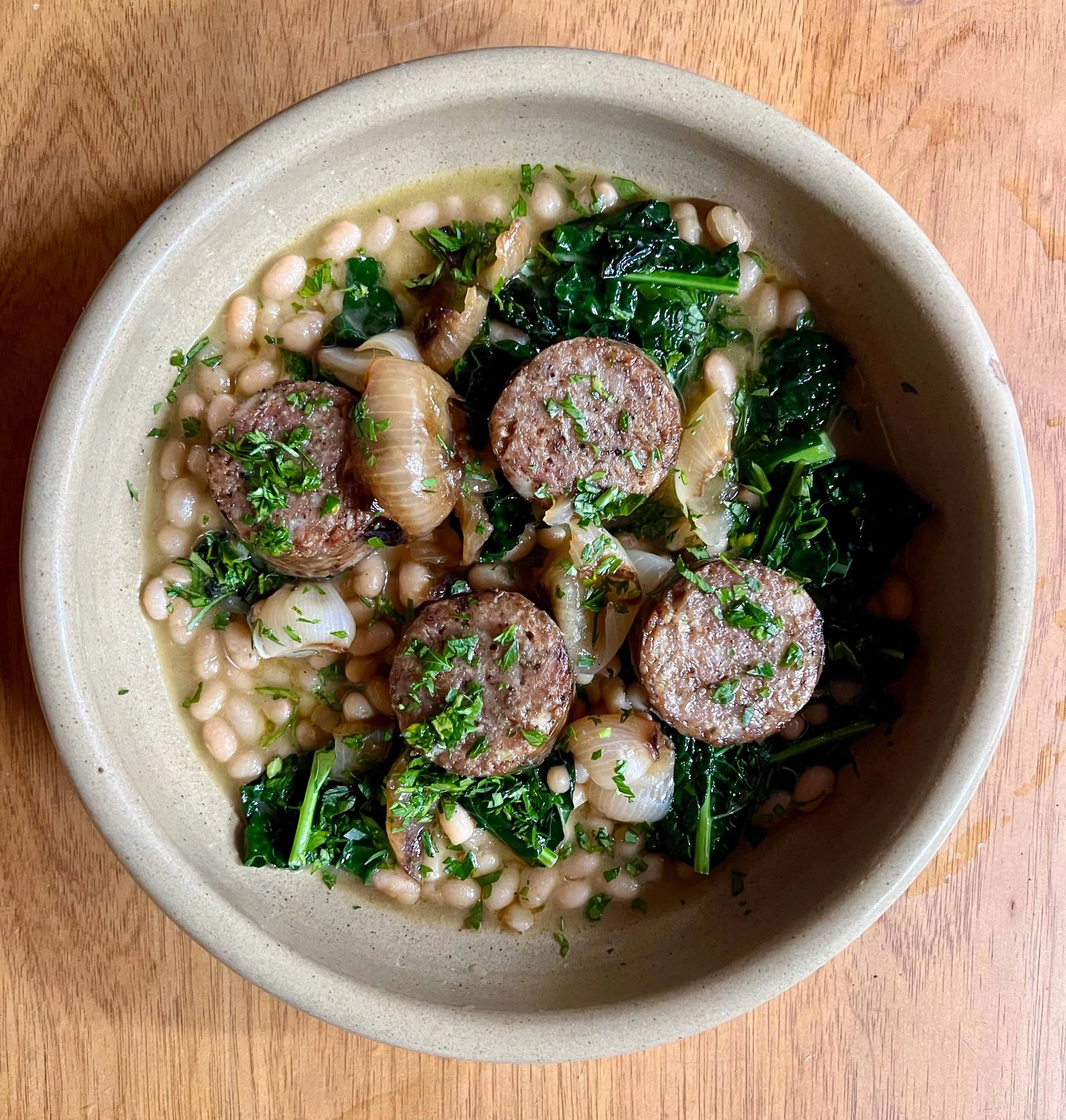 A bowl of white bean soup garnished with chopped parsley, with slices of sausage, onions, and wilted greens, served on a wooden table.